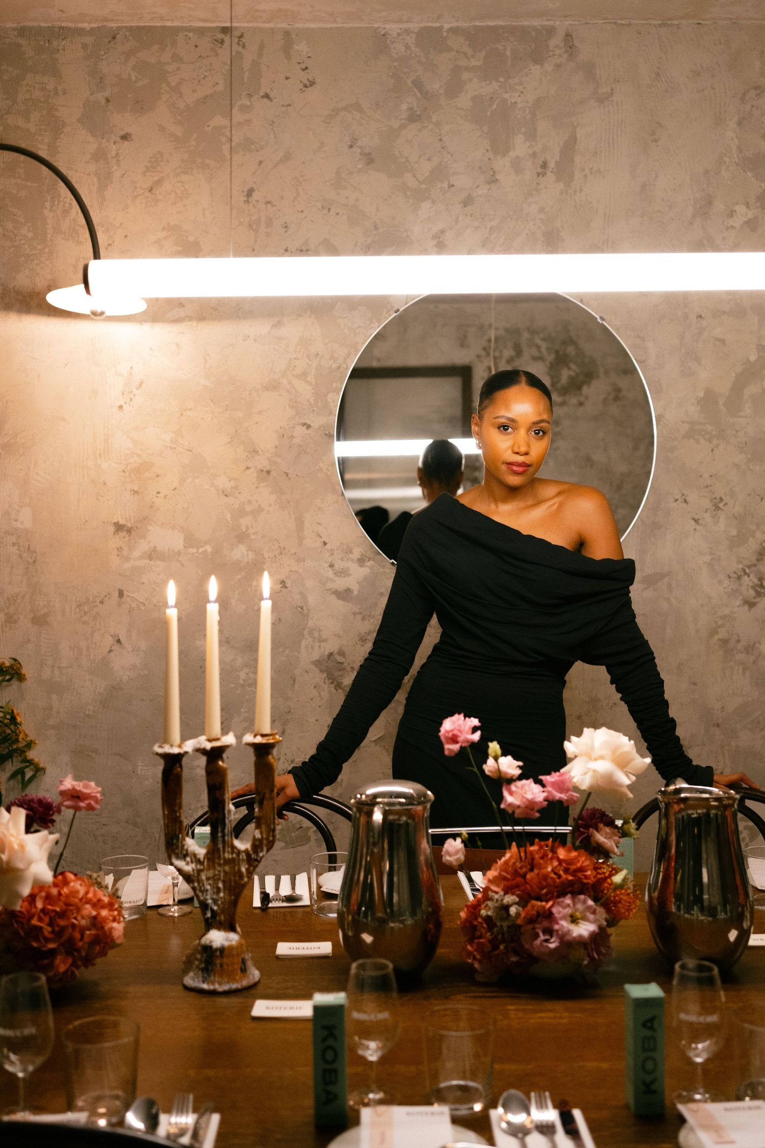 An elegant table setting with white candles, floral arrangements, and crystal glassware against a textured concrete wall.