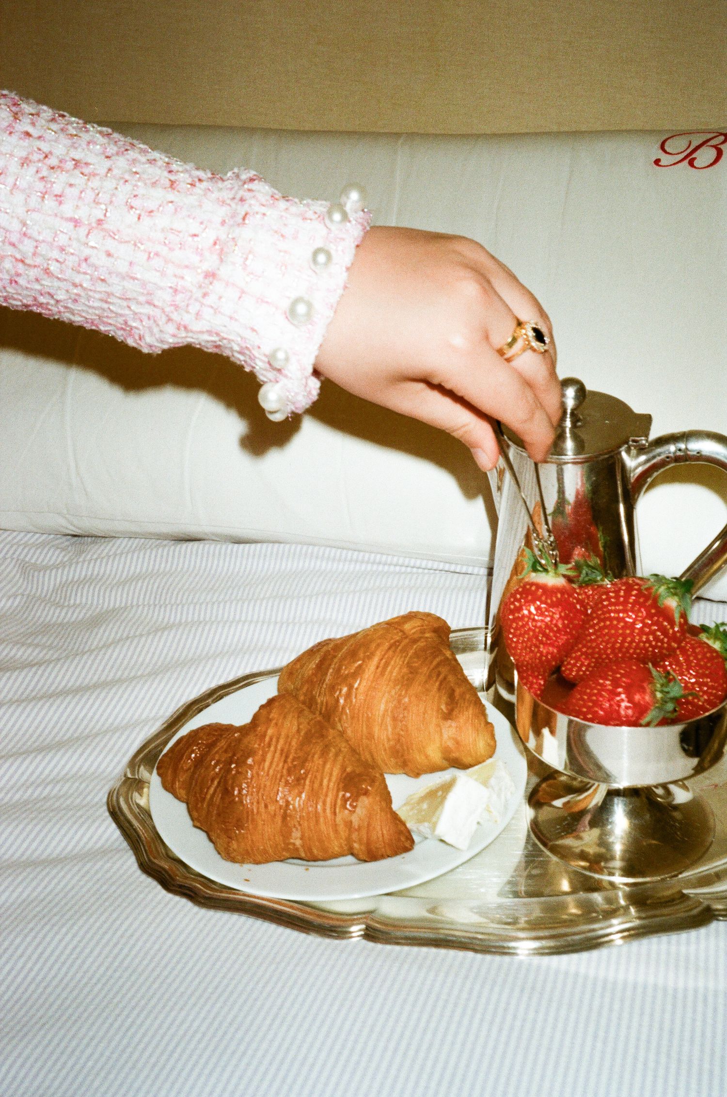 A hand in a lace sleeve reaches for breakfast items including fresh croissants and strawberries on a silver serving tray.
