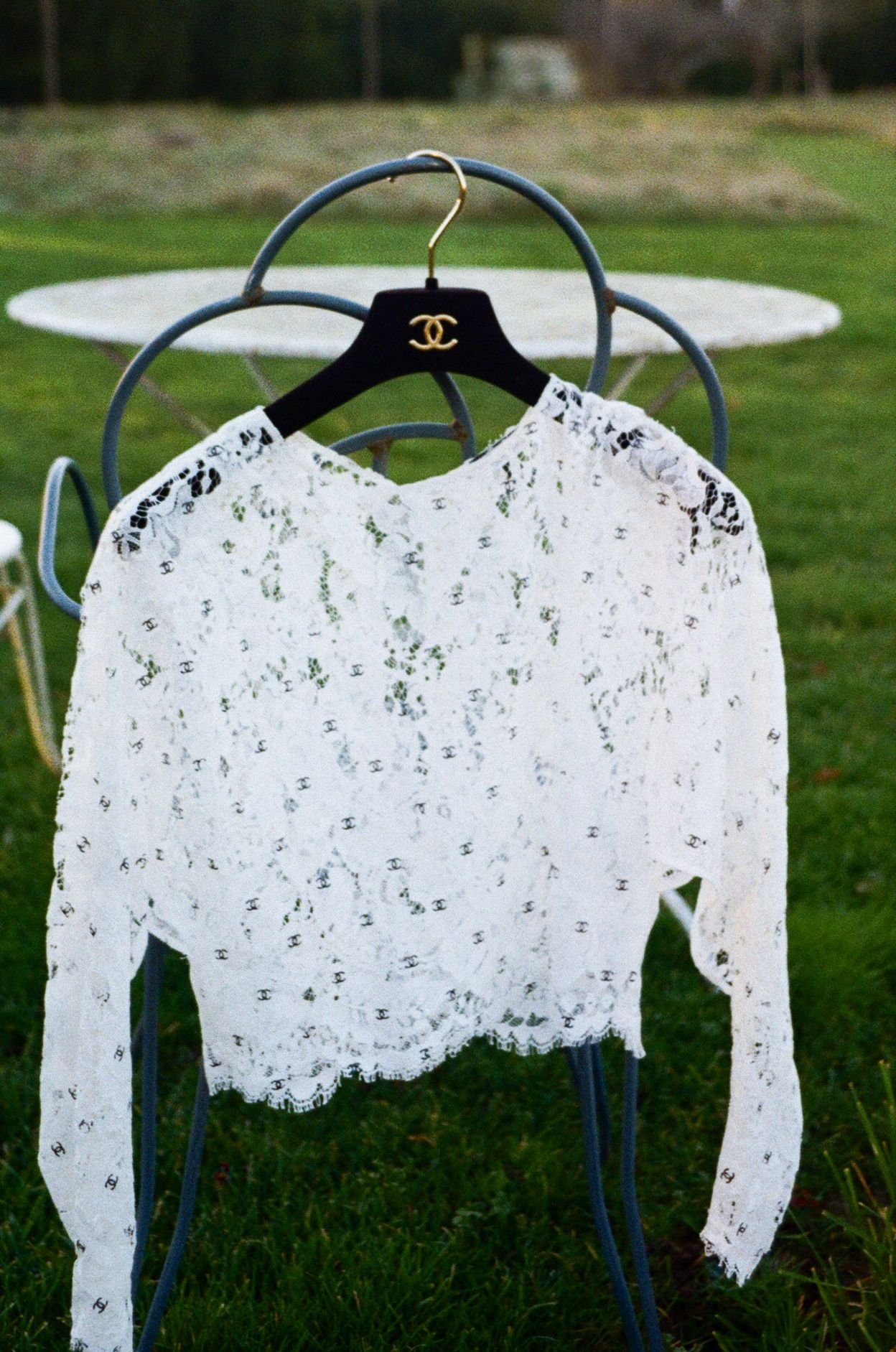 A delicate white lace long-sleeve top hanging on a Chanel hanger against an outdoor garden background.