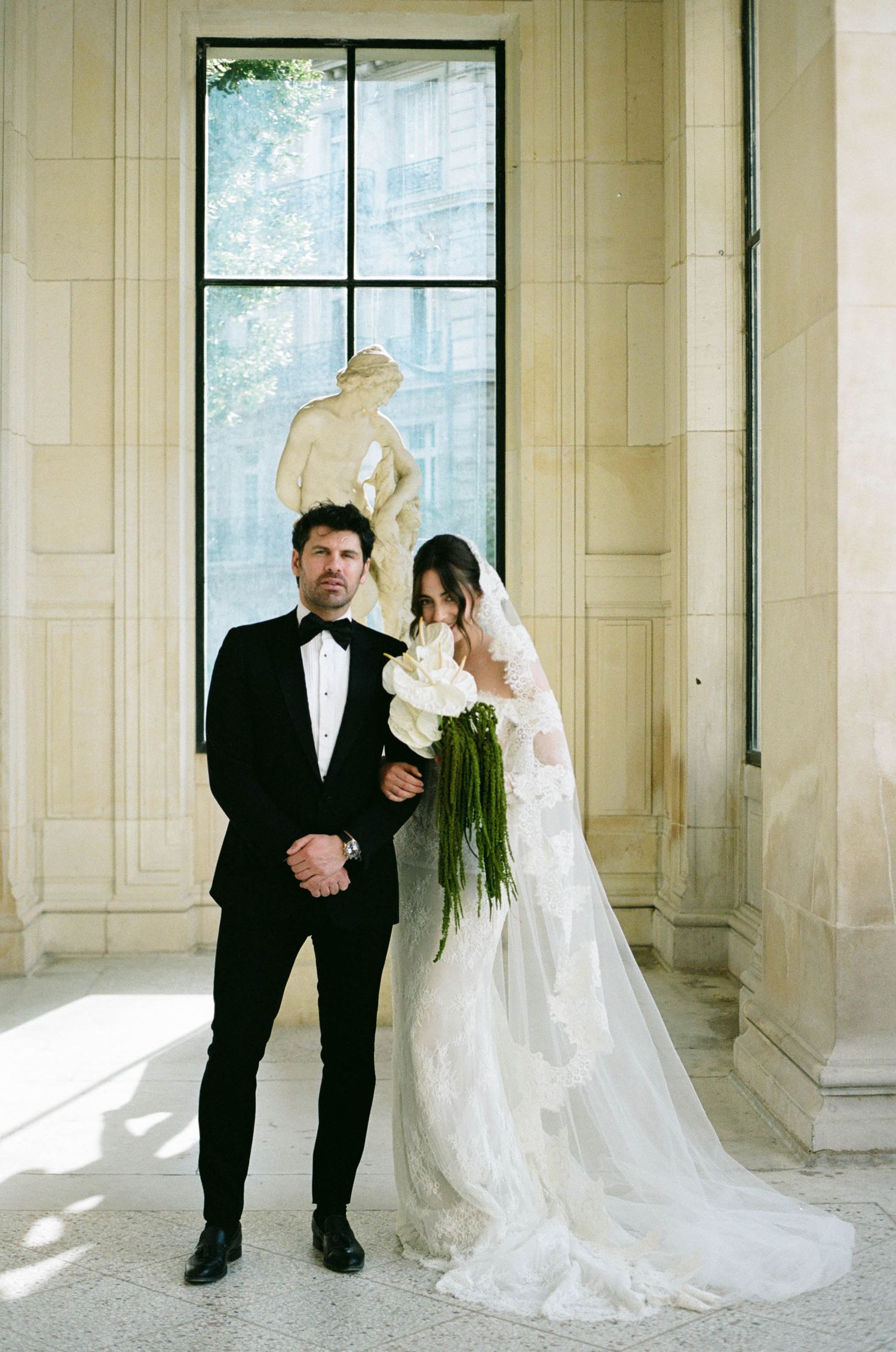 A couple poses for a wedding photo in front of a tall window, with the bride wearing an elegant white gown.