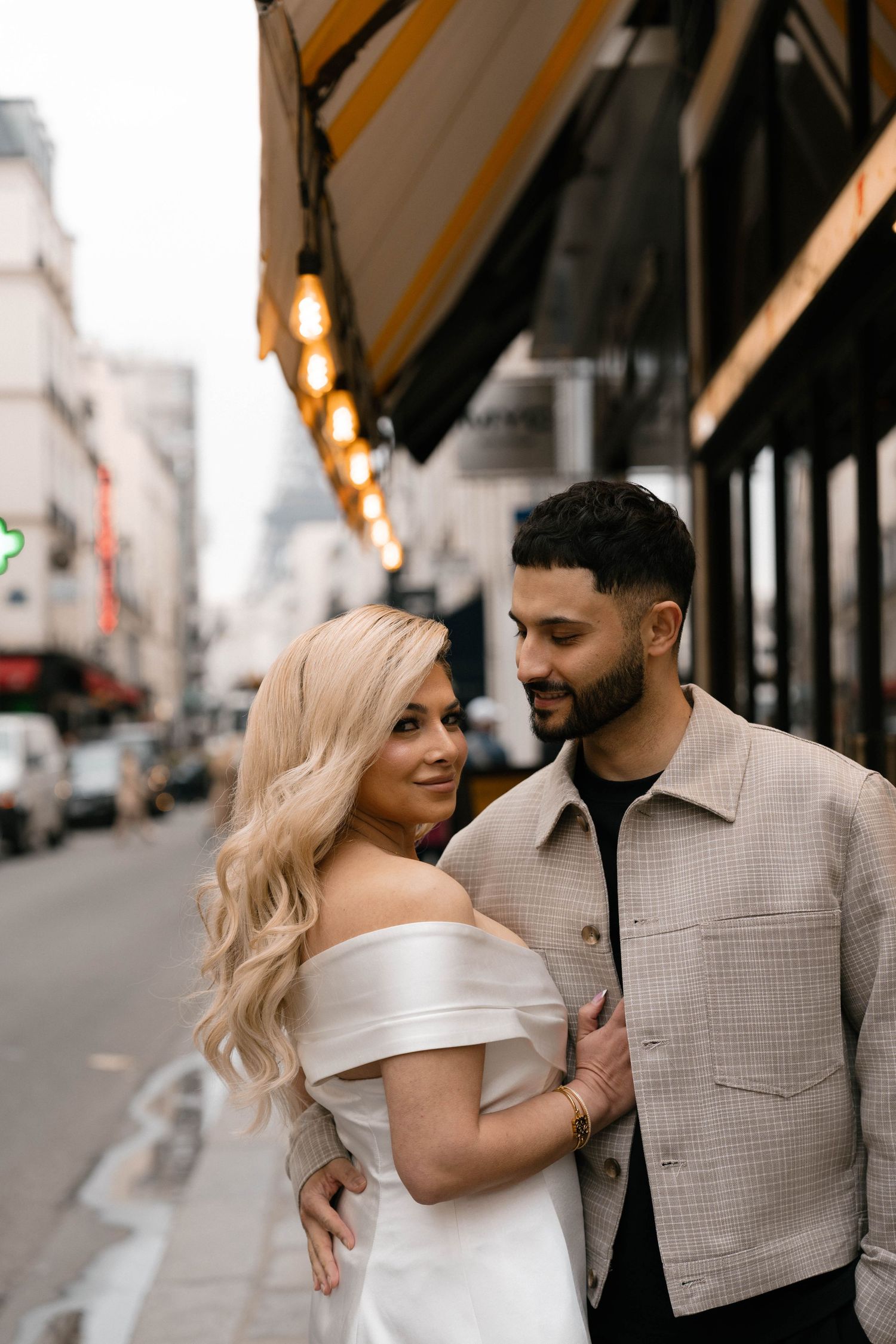 A couple shares loving glances and embraces while walking along a city street with warm lighting and storefronts. PAris film photographer.