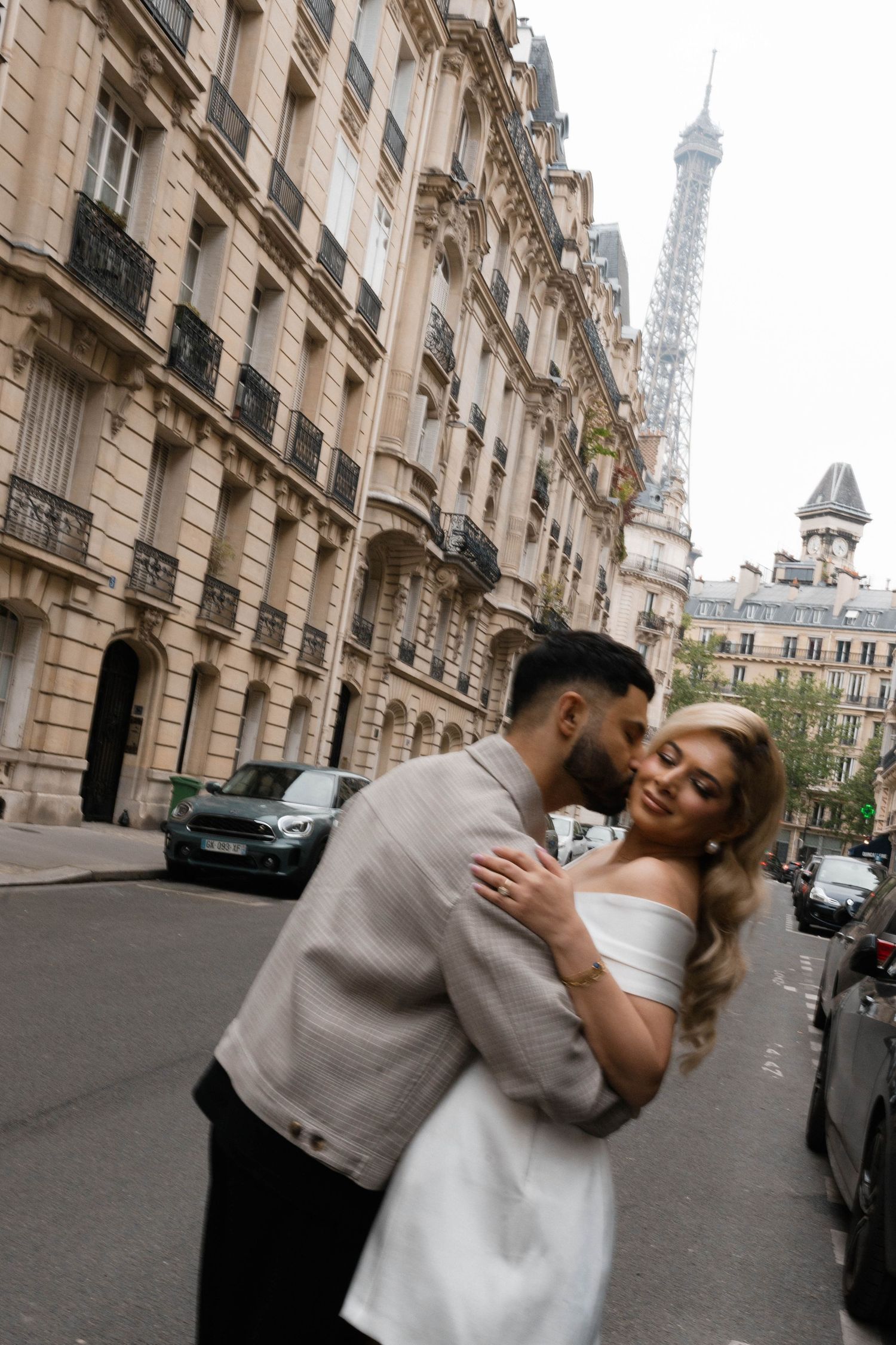 A romantic couple embraces on a Parisian street with the iconic Eiffel Tower visible in the background.