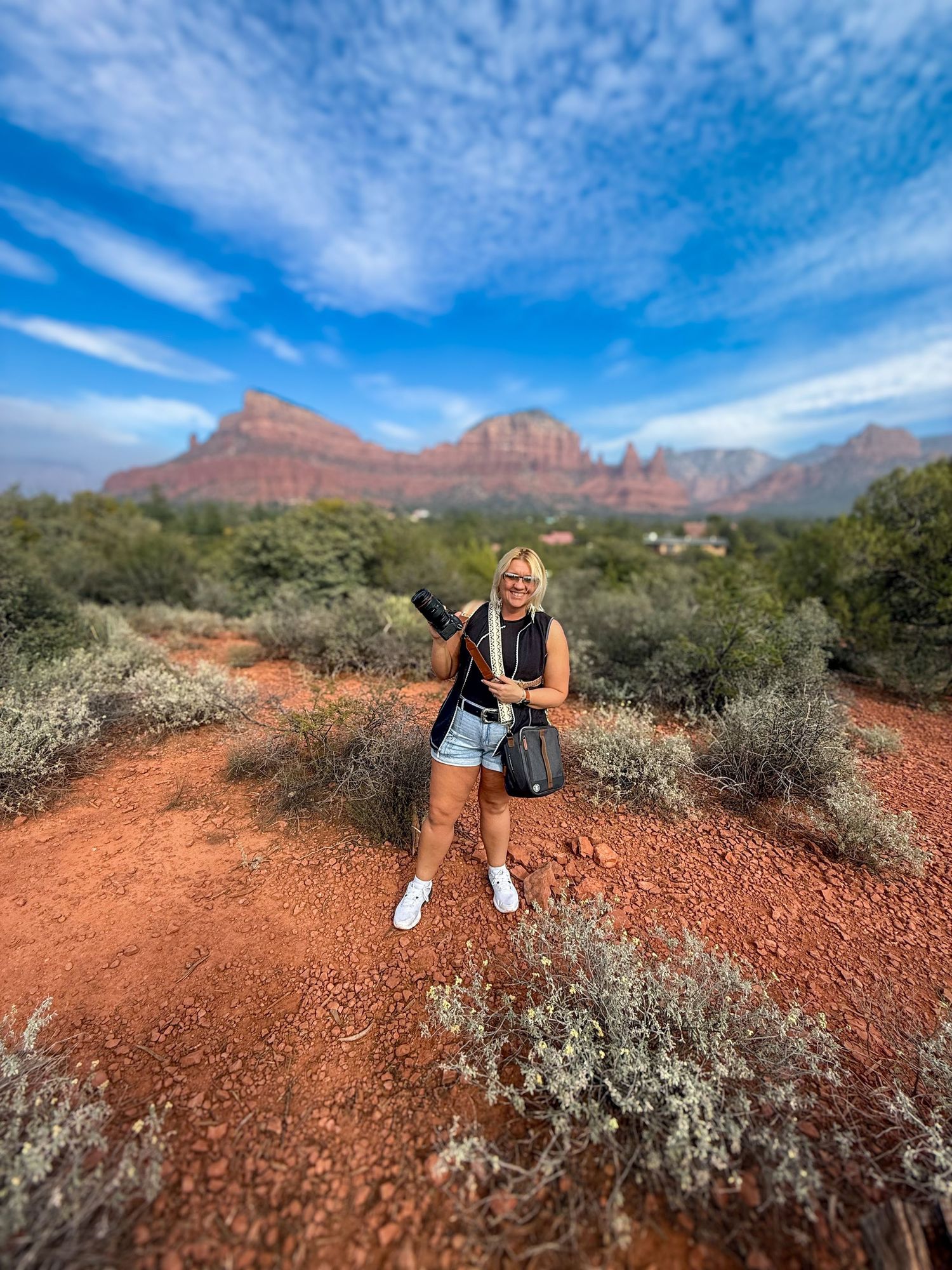 A scenic desert hiking trail with red rocks and mountains of Sedona under bright blue skies with wispy clouds.
