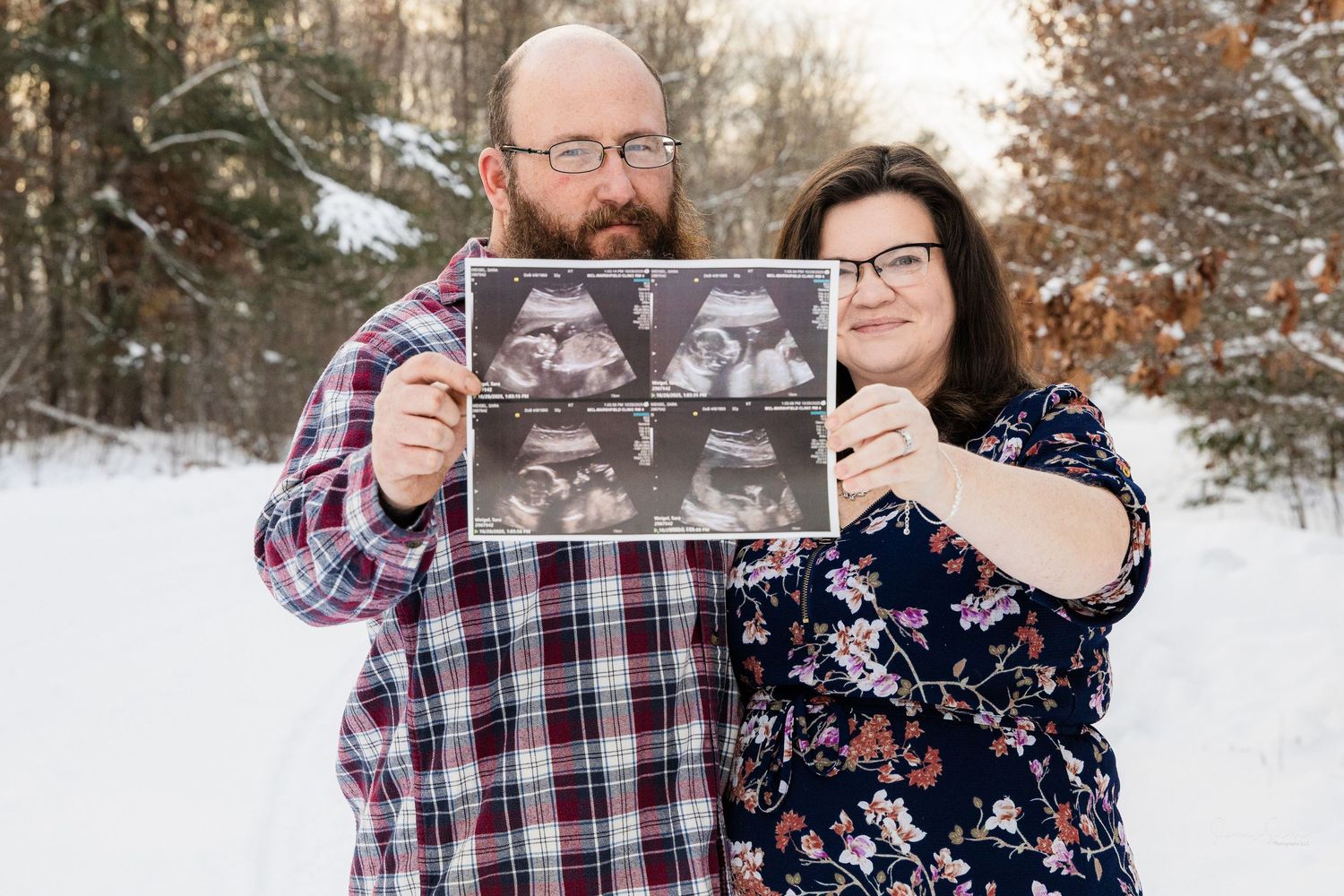 A couple stands in the snow holding up a framed set of ultrasound photos during a winter photo session.