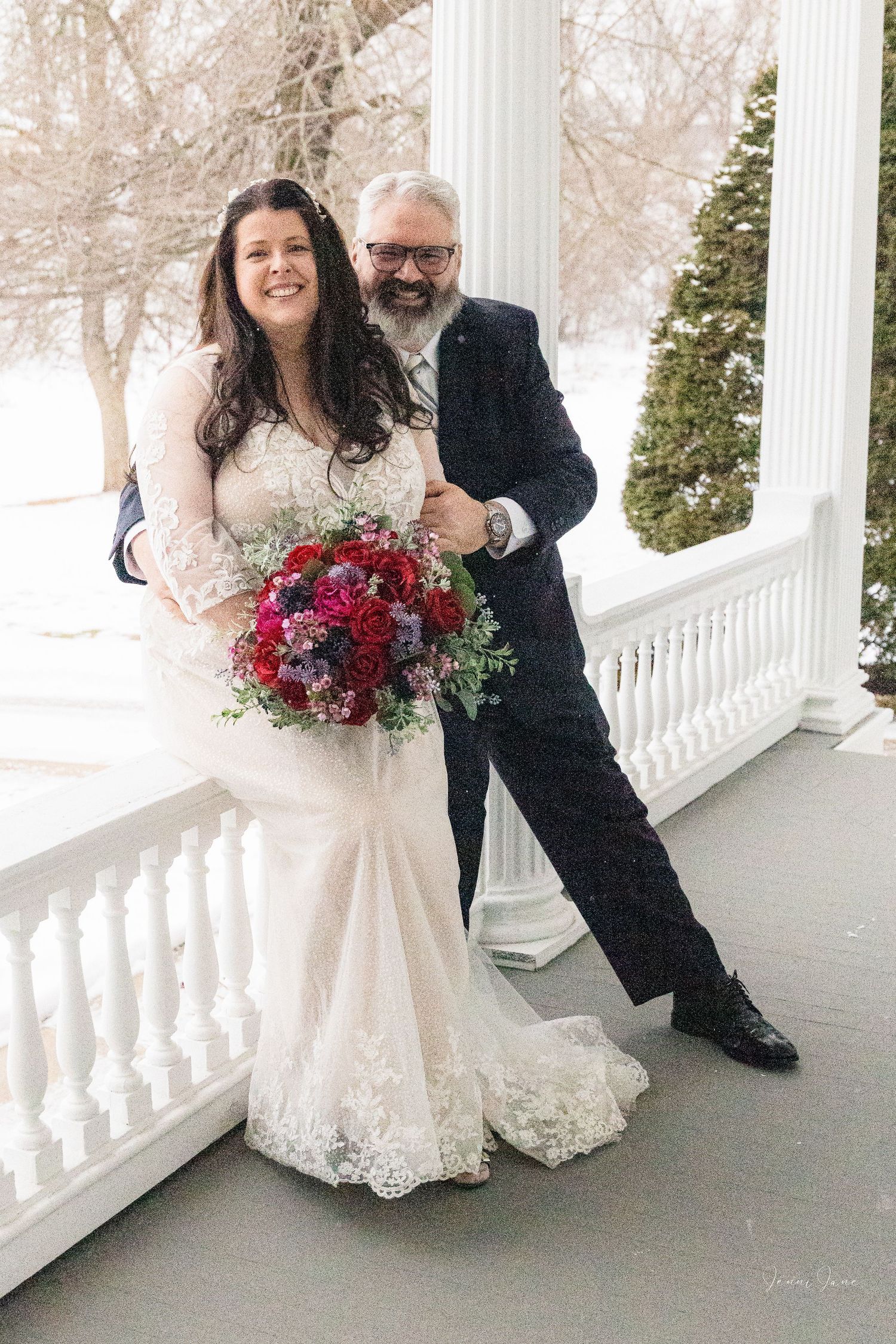 A winter wedding couple poses on a snowy porch with white columns, the bride holding a vibrant red and pink bouquet.