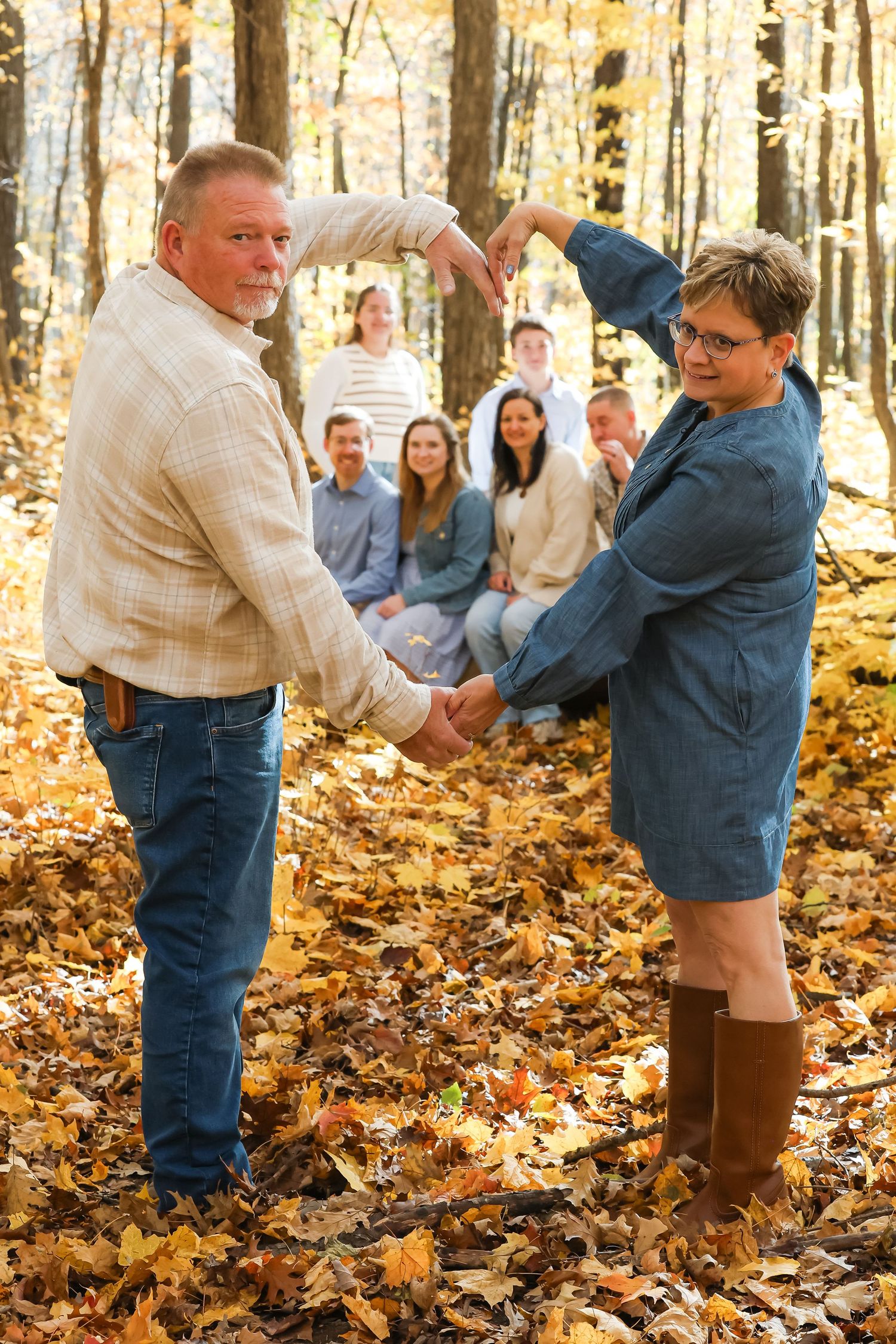 A couple poses together in an autumn forest with fallen leaves while others sit in the background.