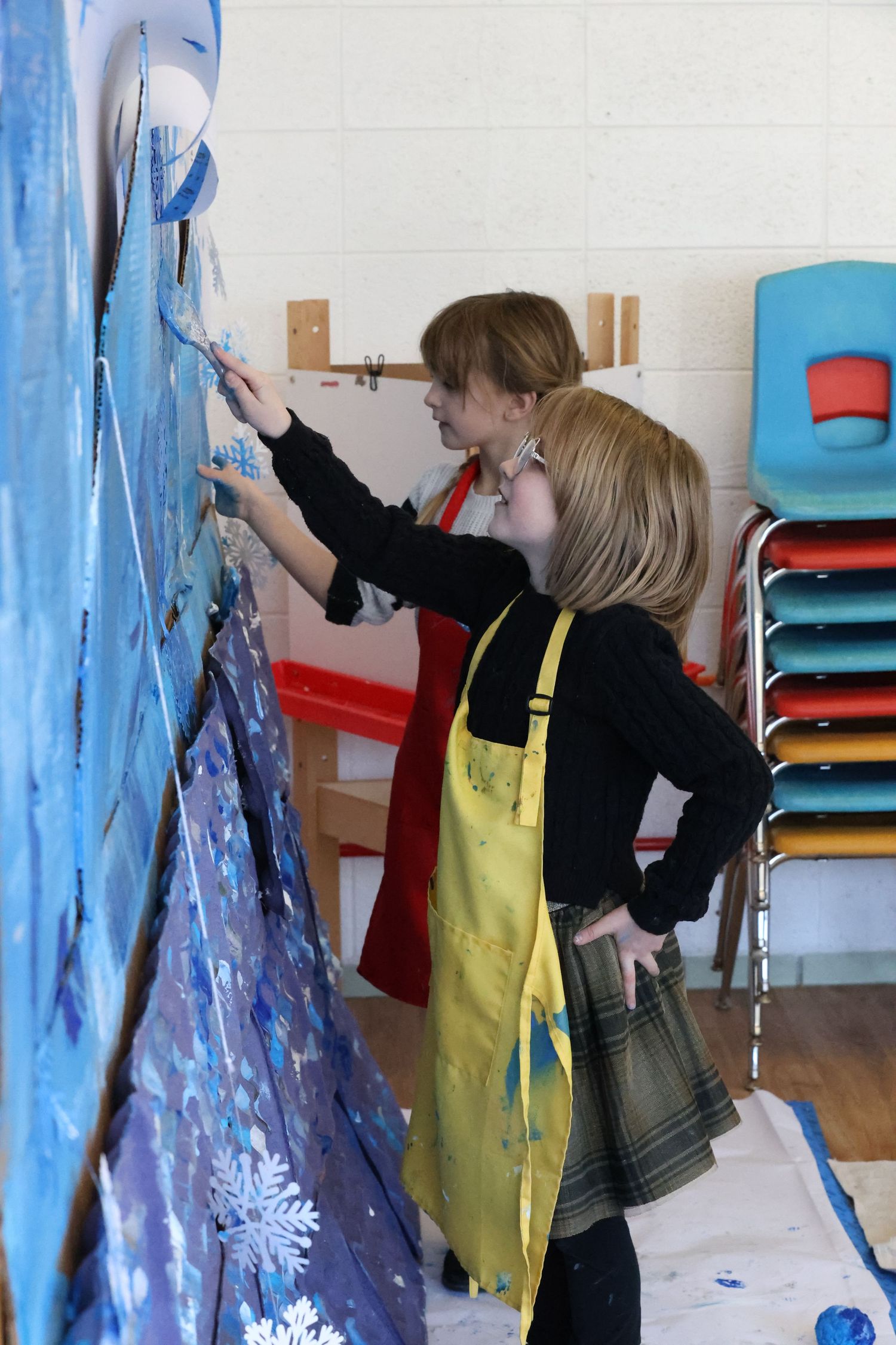 Two artists in yellow aprons paint a large blue mural on a wall while standing near stacked colorful chairs.