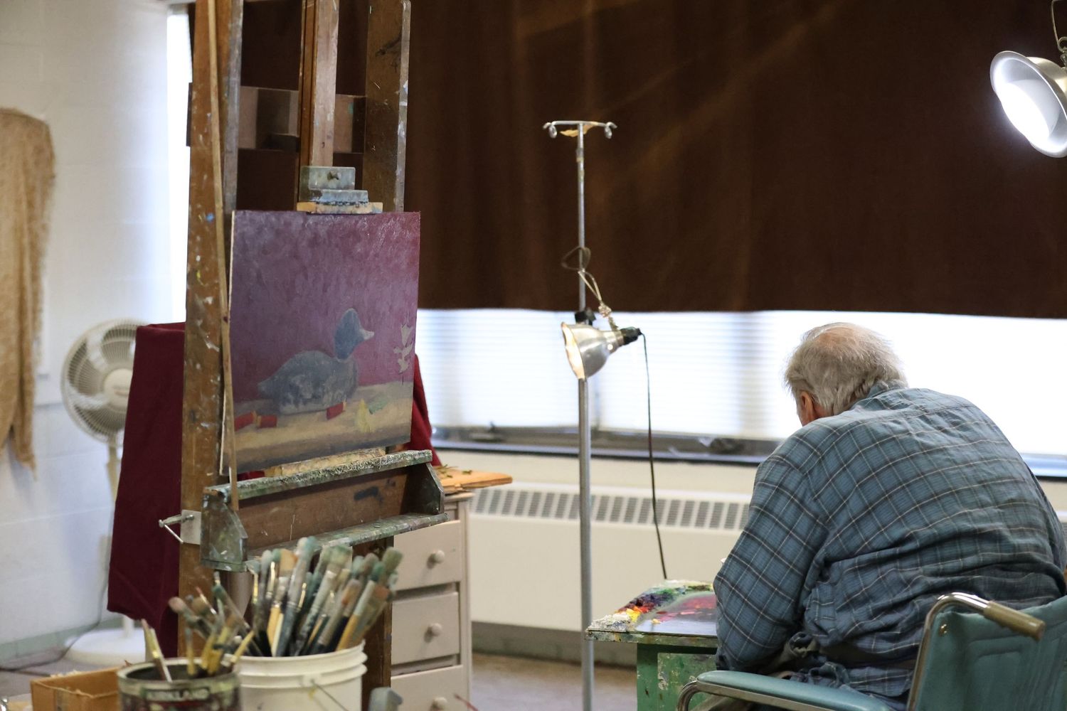 Artist working at an easel in a studio space with painting supplies and an IV stand nearby.