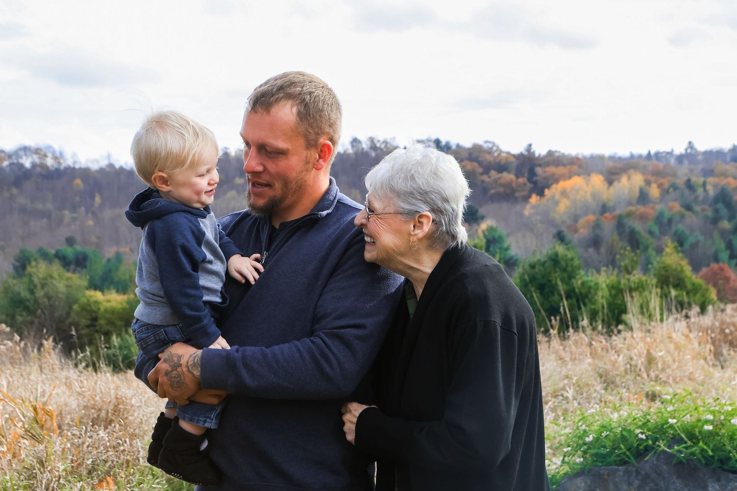 Three generations gather together on a hillside with autumn foliage visible in the background on a sunny day.