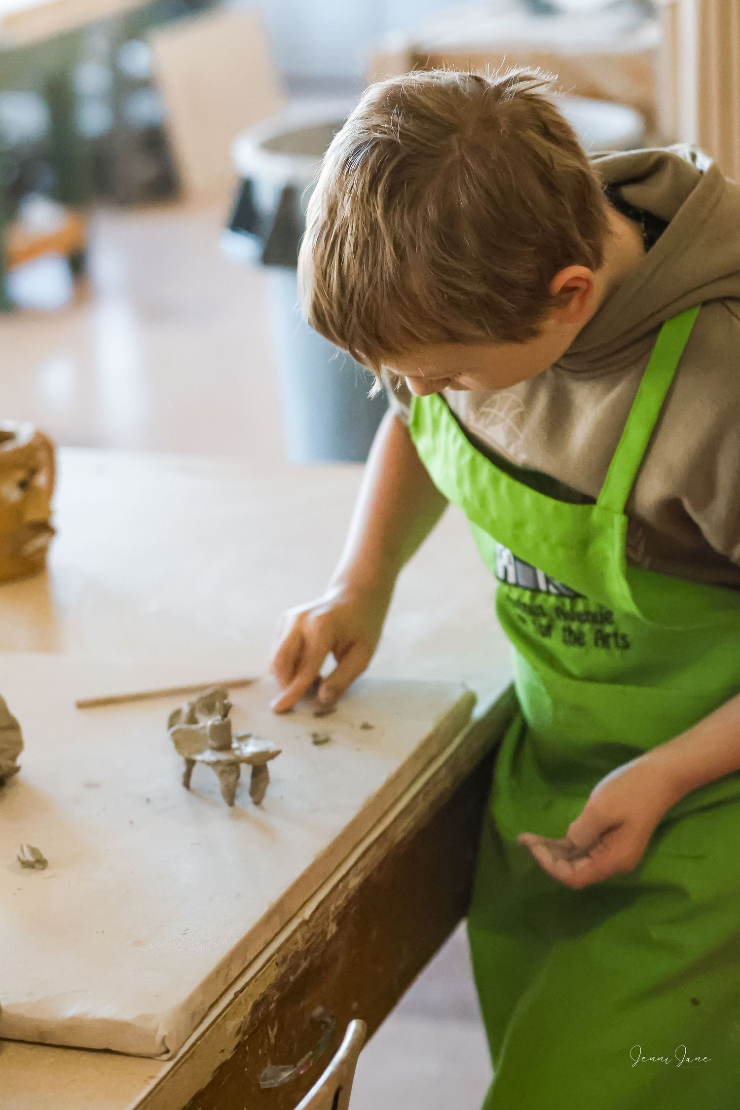 A child in a green apron works with clay or pottery materials on a wooden surface during an art activity.