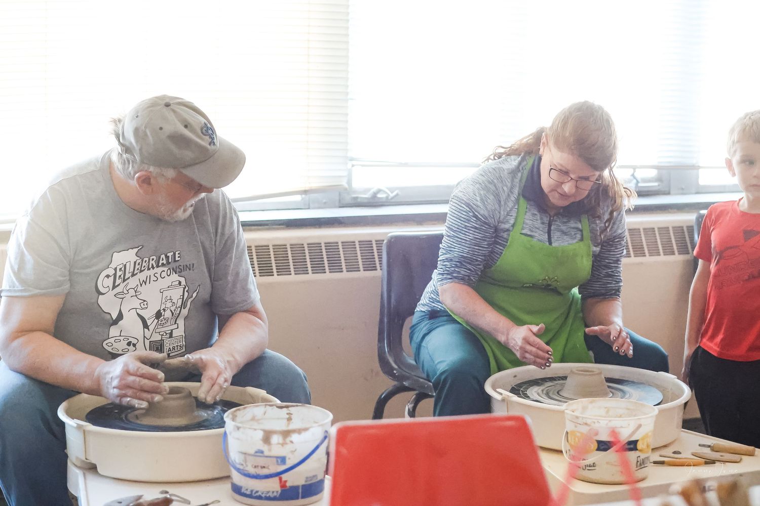 Two people in green aprons work at pottery wheels during a ceramics class in a bright studio space.
