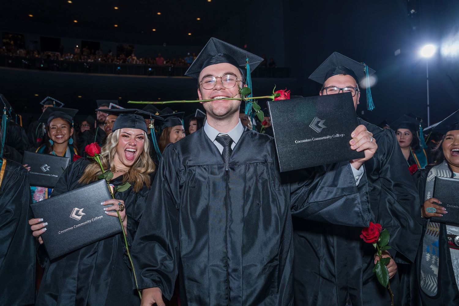 Graduates in black caps and gowns celebrate at commencement ceremony, holding diplomas and roses while smiling.