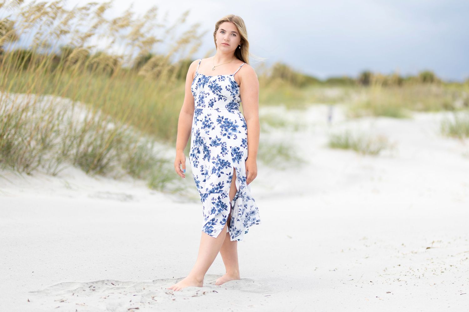 Woman in white floral blue dress standing on sandy beach dunes on a sunny day.