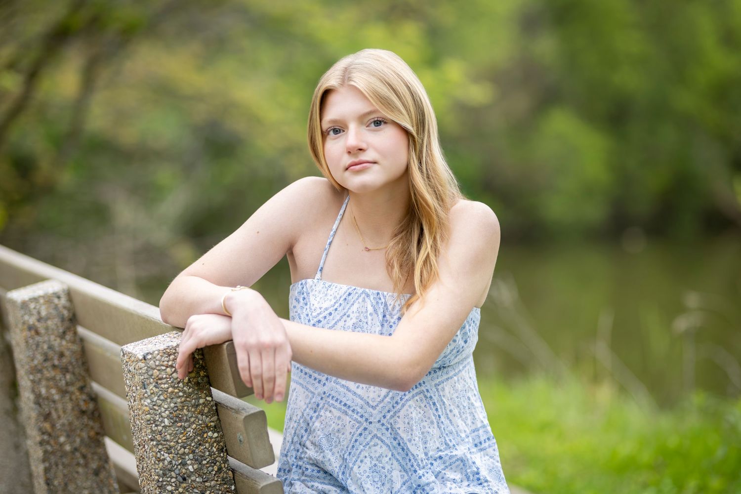 Young blonde woman in blue floral sundress leaning on a railing outdoors near a pond.