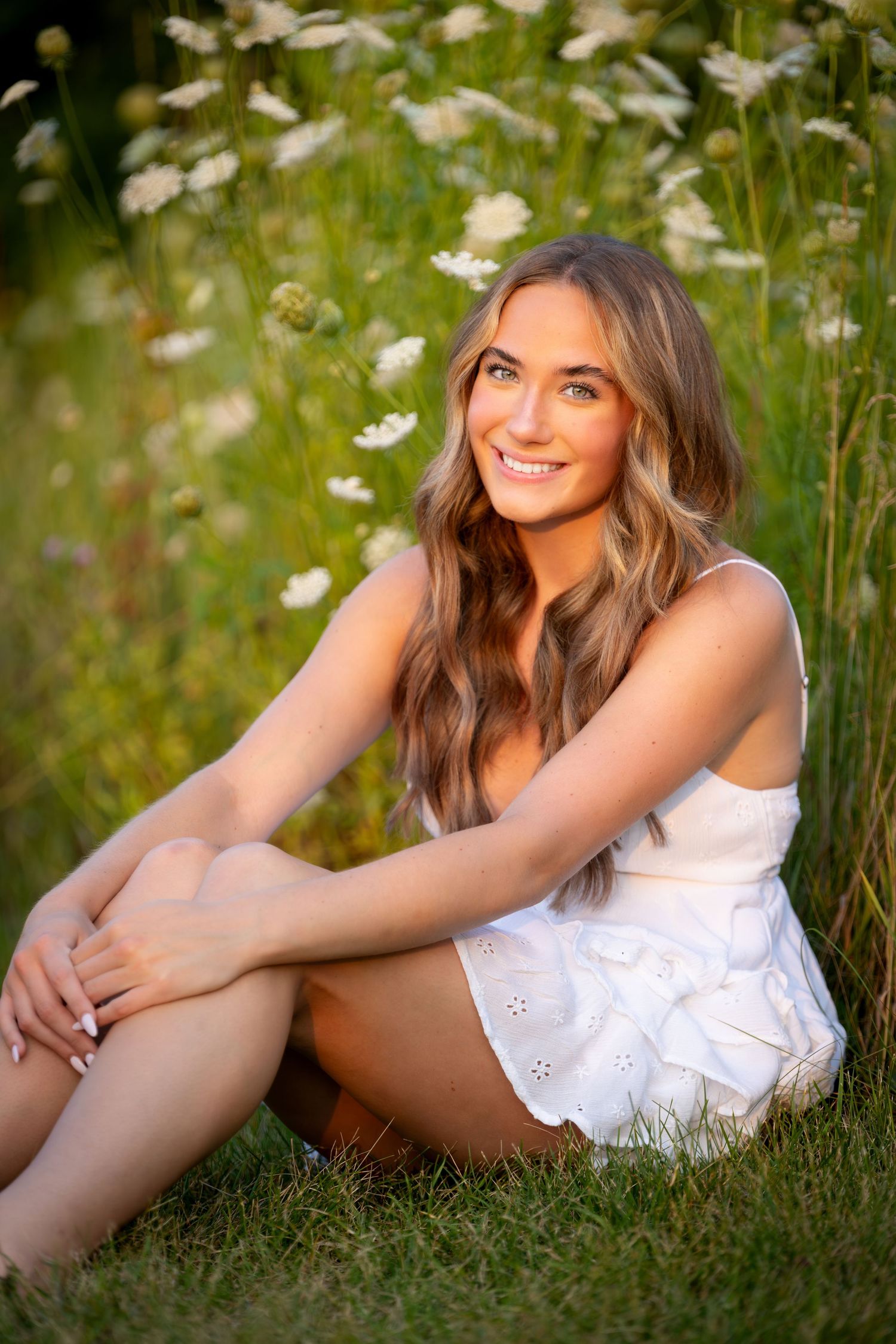 Young woman in white dress sitting in green field with wildflowers, smiling outdoors in warm sunlight.