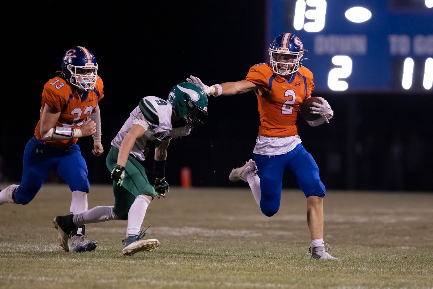 Orange-uniformed football player stiff-arms green defender while running with ball during night game.