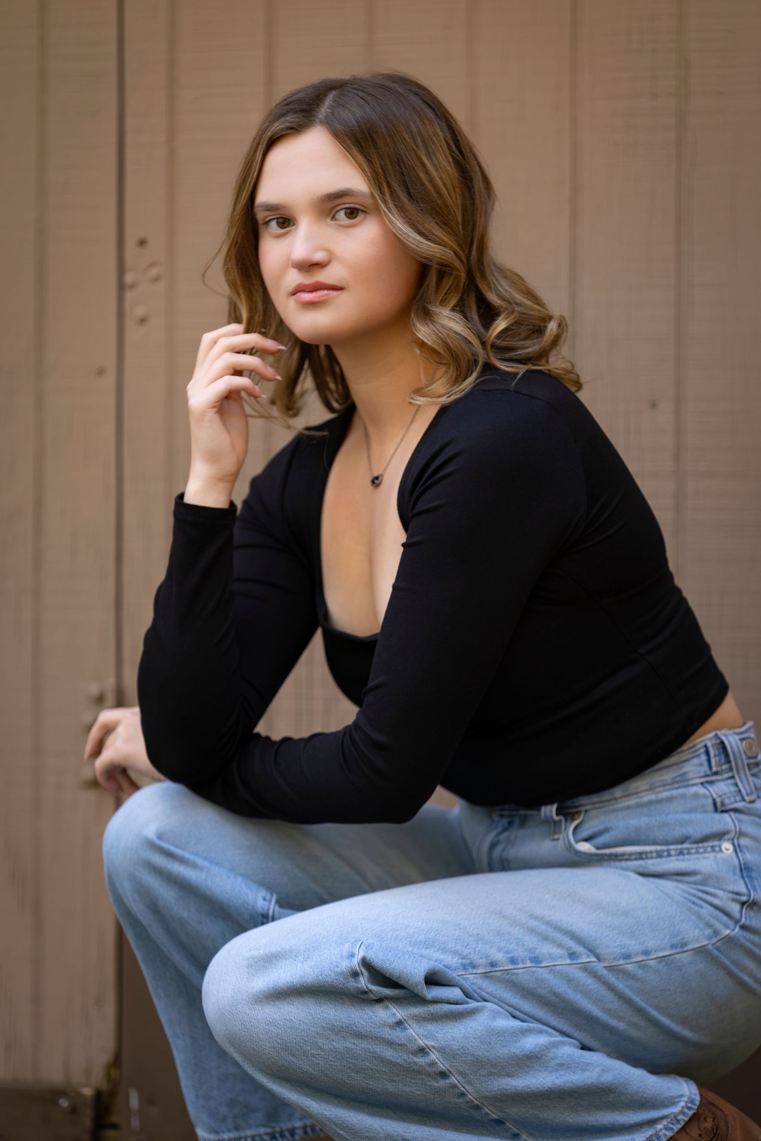Young woman in black long-sleeve top and jeans posing confidently against a wooden backdrop.