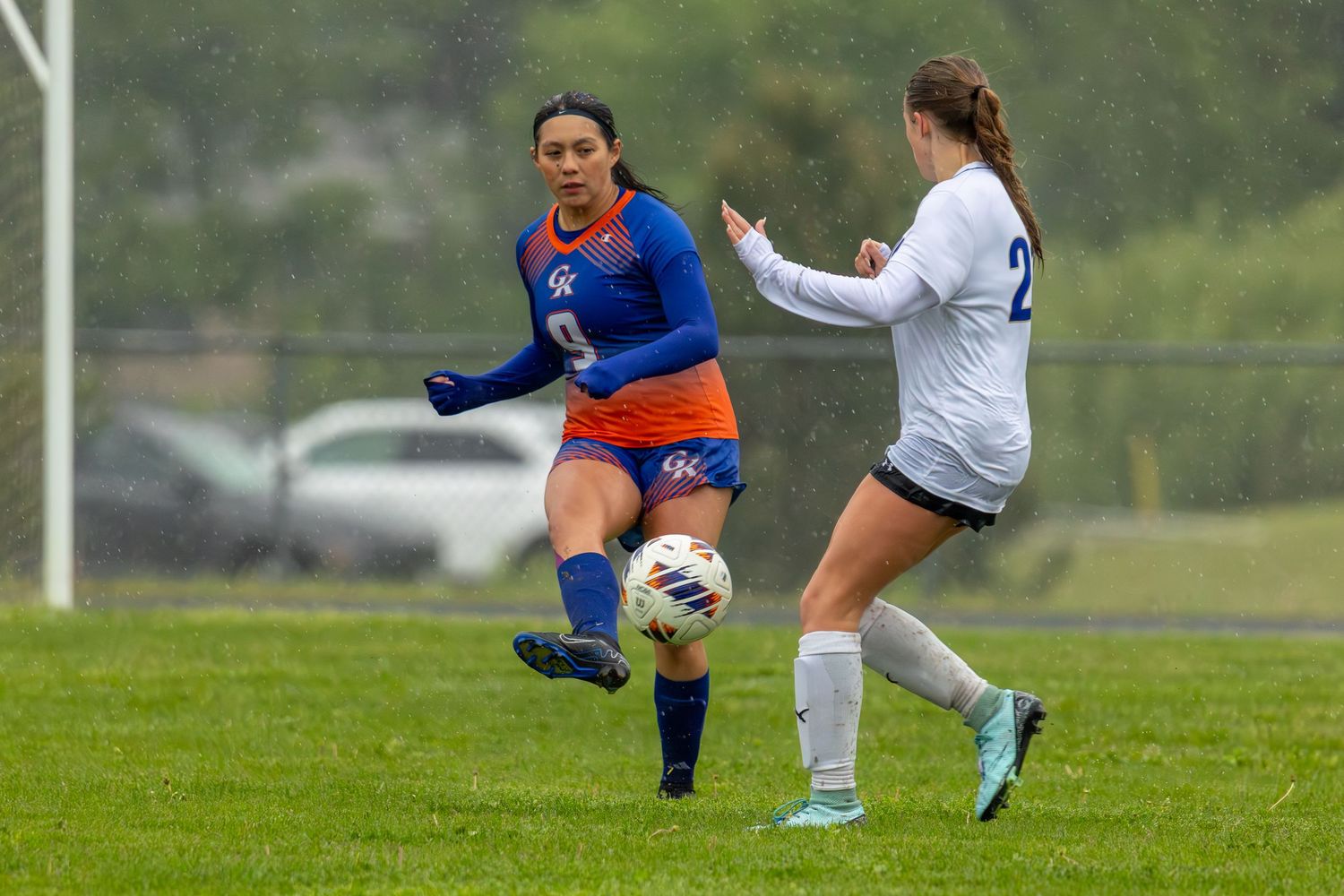 Two female soccer players compete for the ball during a rainy match, one in blue and one in white.