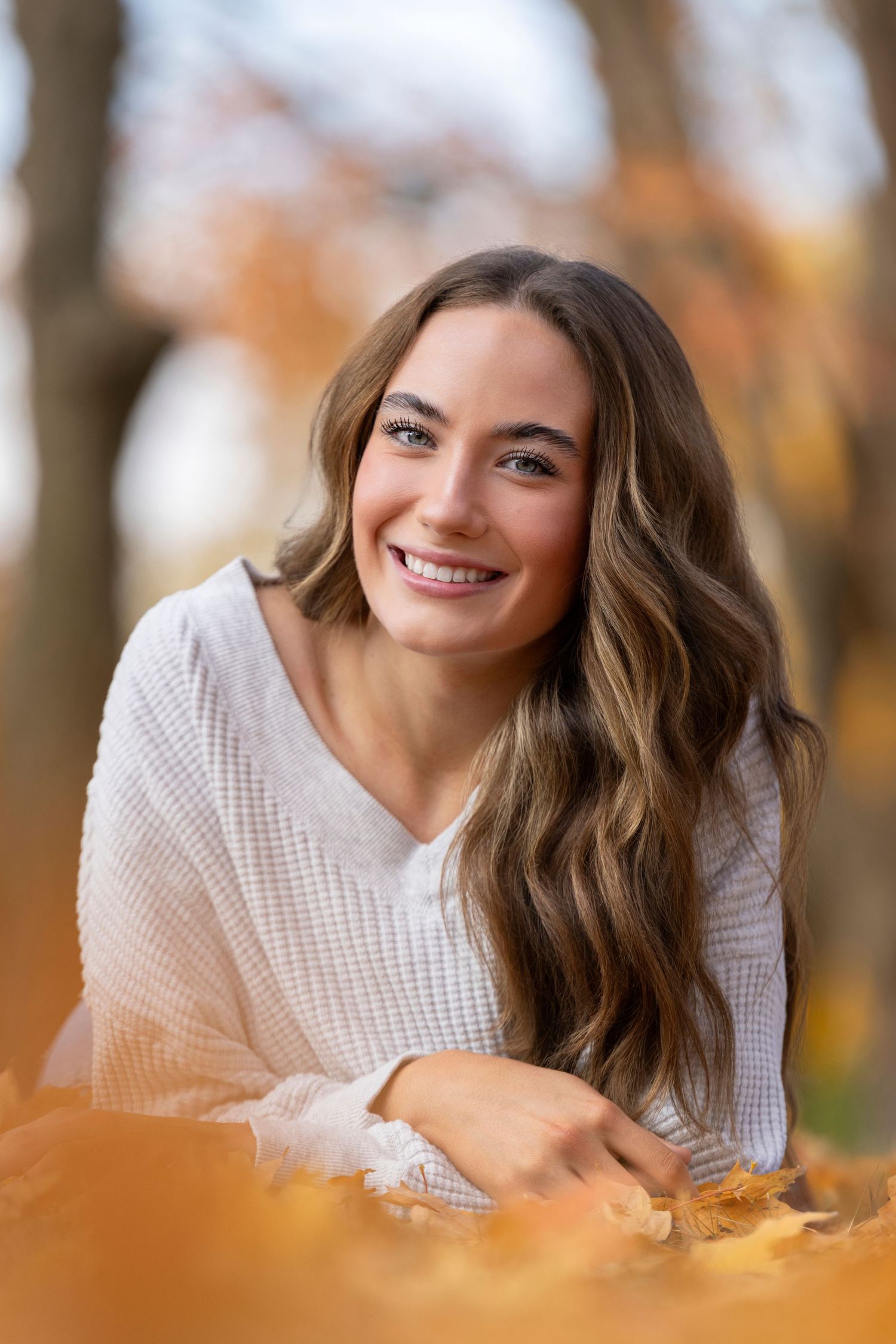 Young woman with long wavy hair smiling among autumn leaves in a park wearing a white sweater.