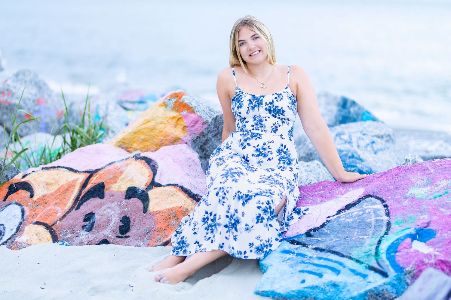 Woman in blue floral maxi dress sitting on sandy beach surrounded by colorful towels, smiling outdoors.