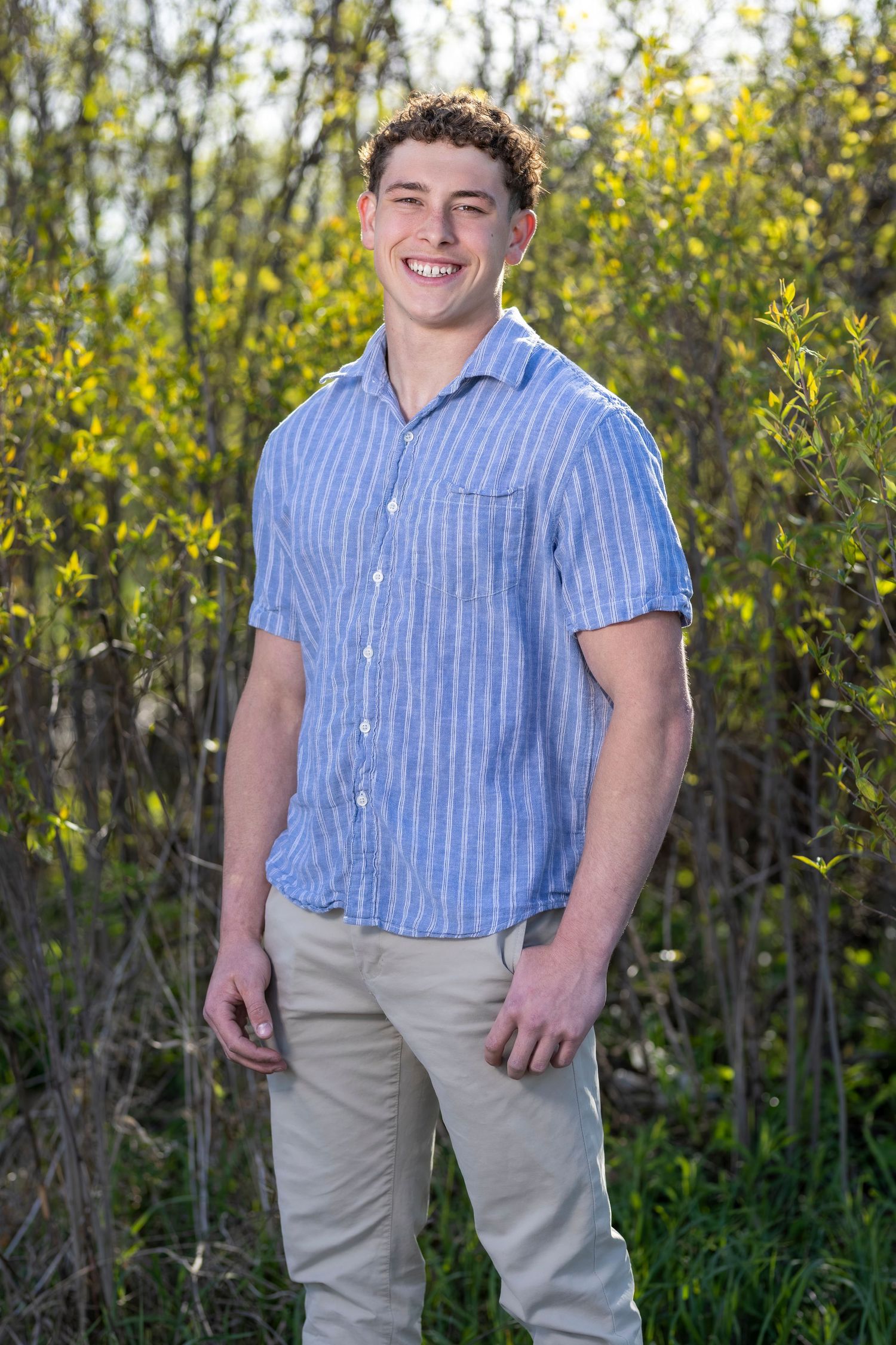 Young man smiling outdoors in blue striped shirt and khaki pants, standing among green spring foliage.