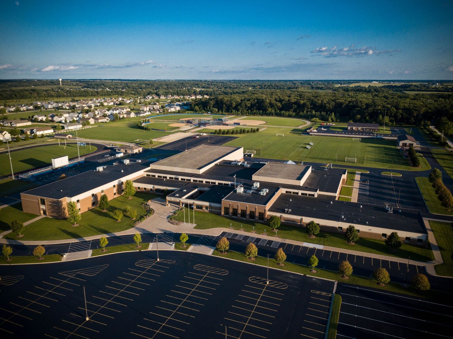 Aerial view of a modern school campus with large parking lot, sports fields, and surrounding suburban landscape.