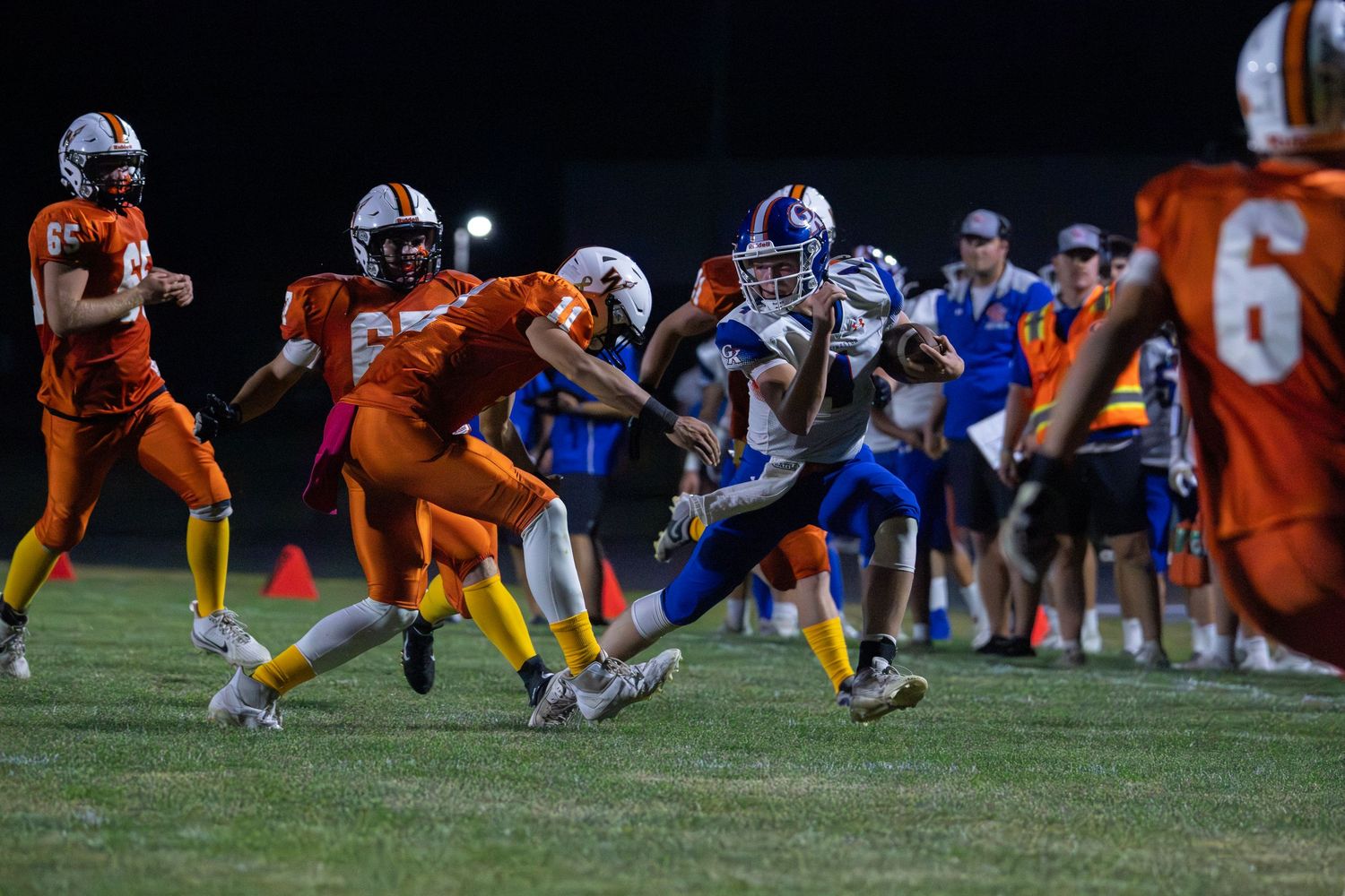 High school football player in blue uniform breaks through tackle attempt by orange-jersey defenders during night game.