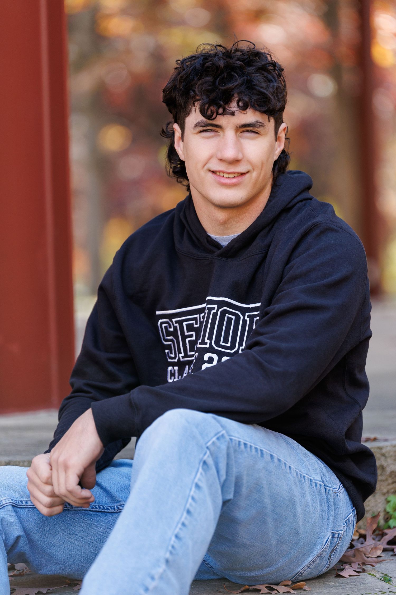 Young man with curly dark hair sits outdoors in a black hoodie and jeans, smiling against autumn foliage backdrop.