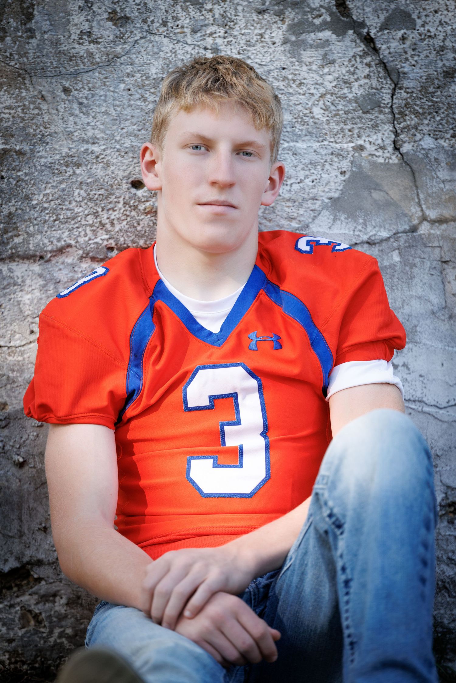 Young male athlete in orange number 3 football jersey sitting against stone wall for senior portrait photo.