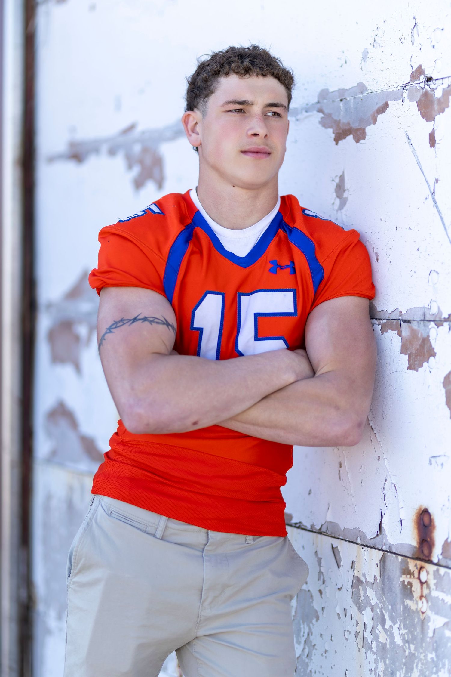 Young athlete in orange Under Armour football jersey number 15 with arms crossed leaning against weathered white wall.