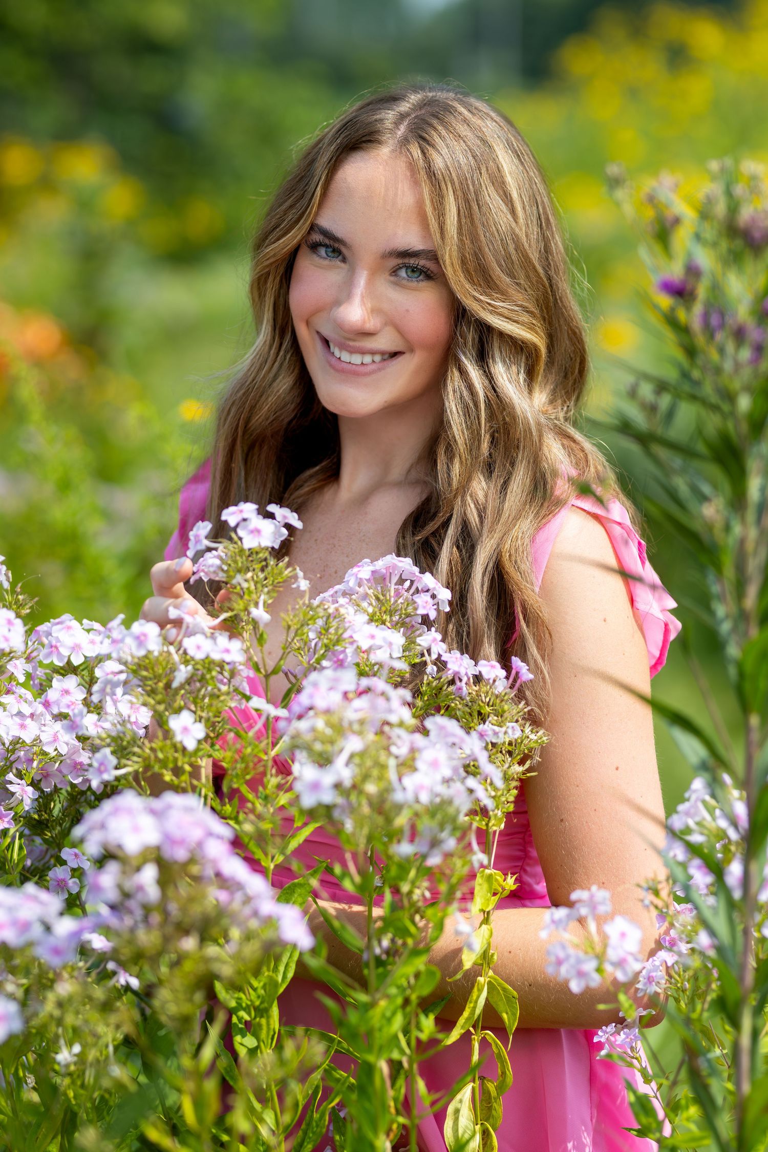 Smiling young woman in pink dress holding purple wildflowers in a lush garden.