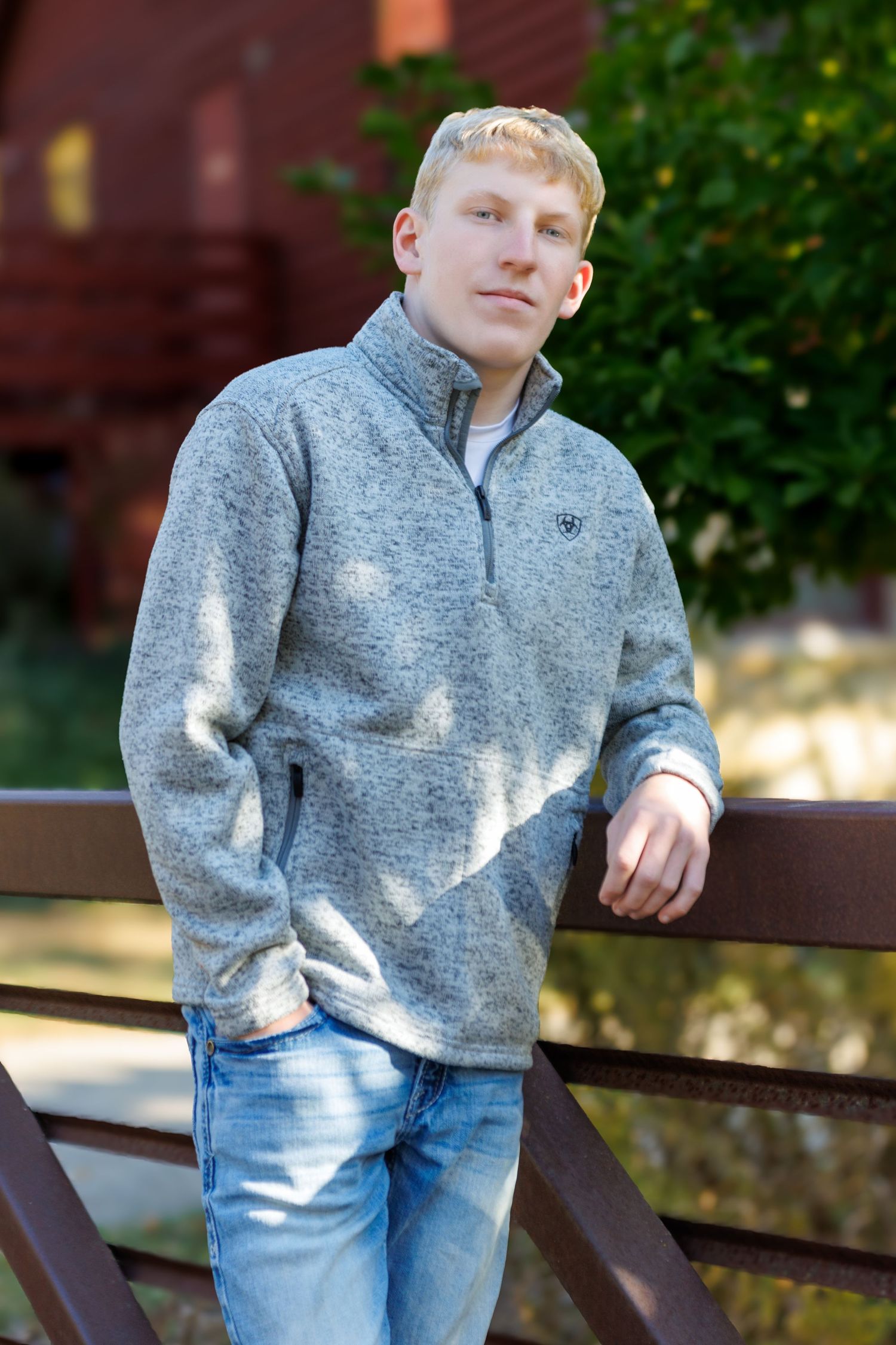 Young man in gray Ariat quarter-zip fleece and jeans leaning on rustic fence for outdoor senior portrait.