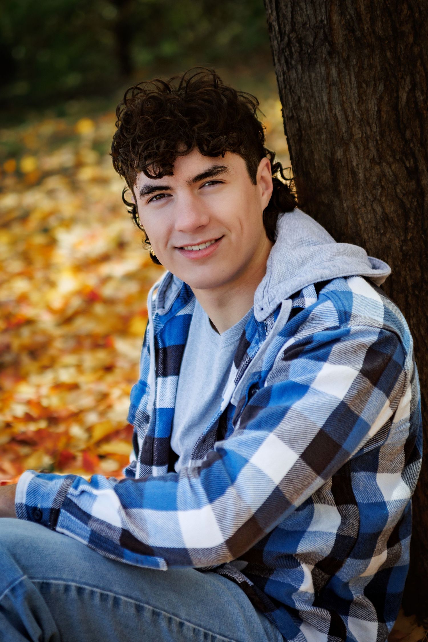 Smiling young man with curly dark hair leaning against a tree in autumn park, wearing blue plaid flannel shirt.