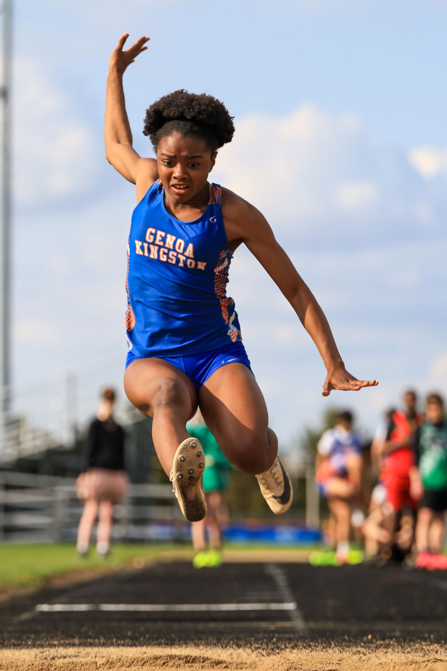 Genoa Kingston athlete performs long jump mid-air during outdoor track and field competition in blue uniform.
