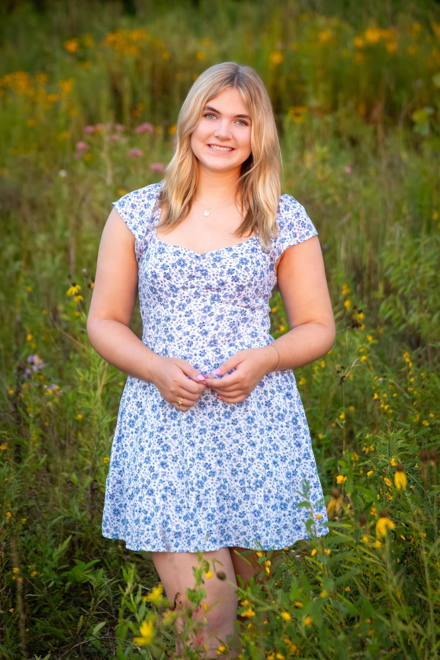 Young blonde woman in a blue floral dress smiling in a wildflower meadow during golden hour.