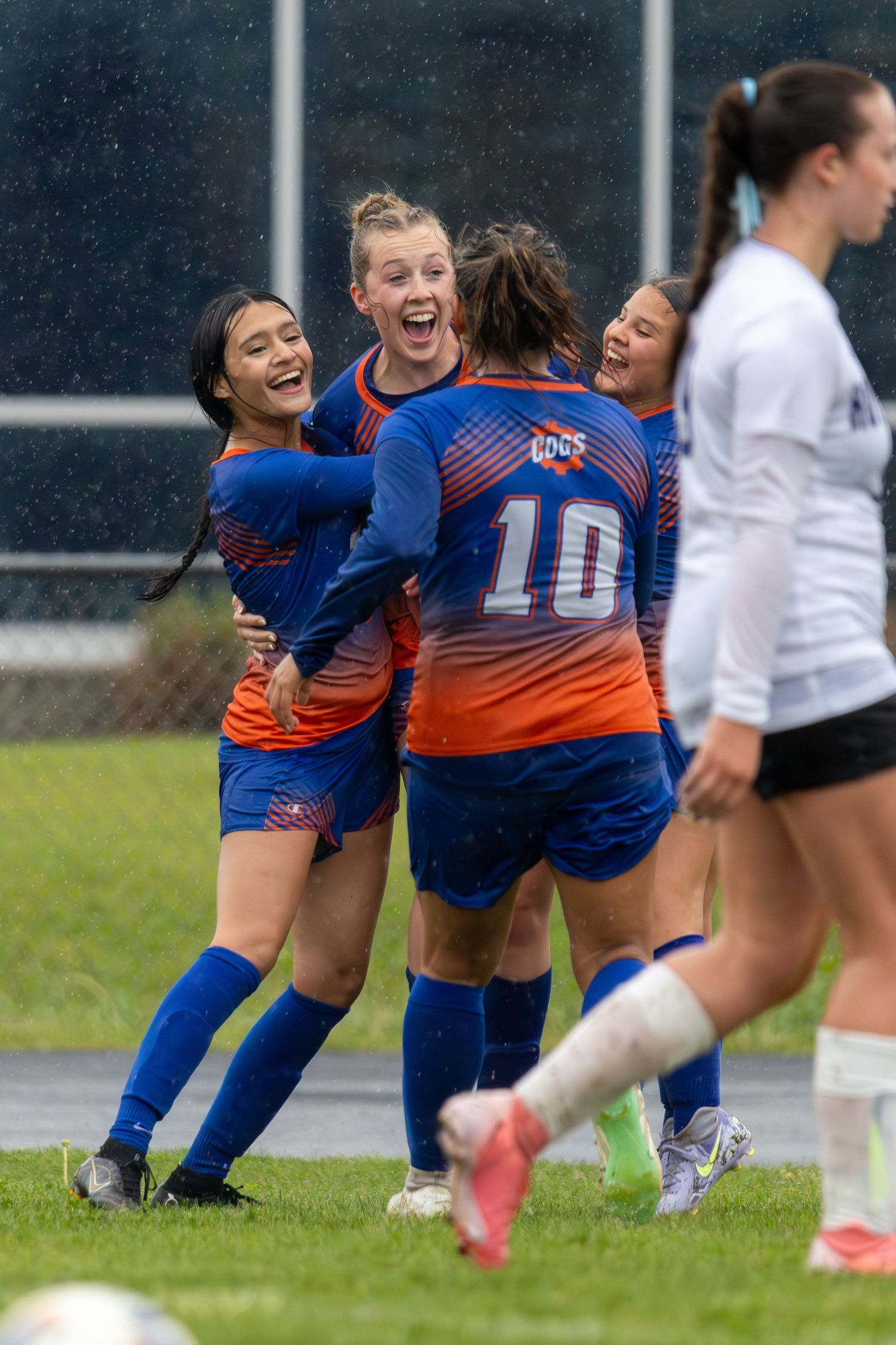 Girls soccer team in blue and orange uniforms celebrating together on the field during a rainy match.
