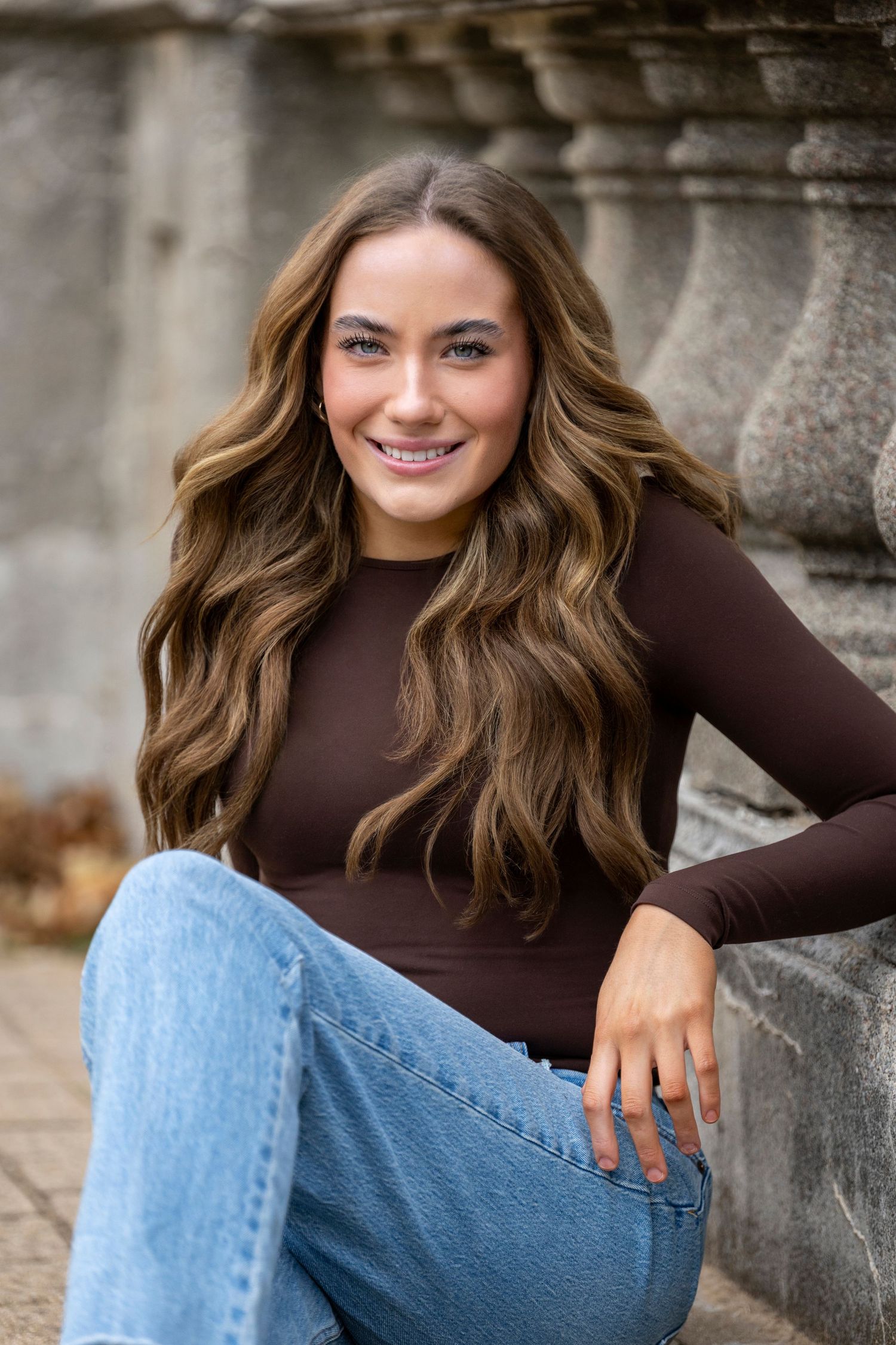 Young woman with long wavy brown hair smiles in a brown top and jeans, seated outdoors near stone columns.