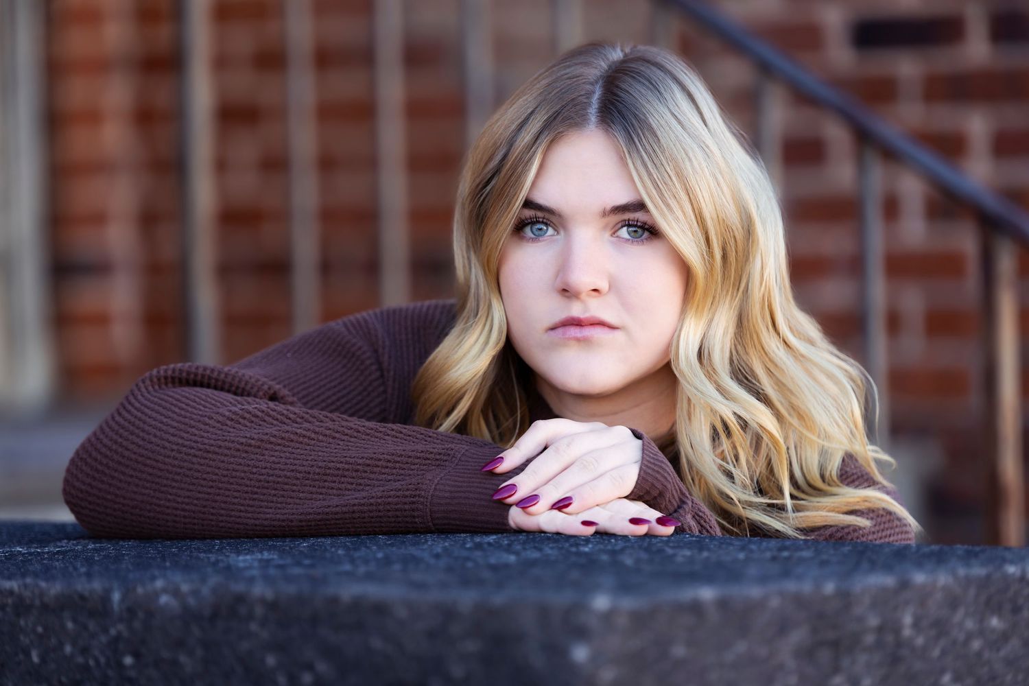 Young blonde woman resting chin on hands, posing outdoors against brick wall background.