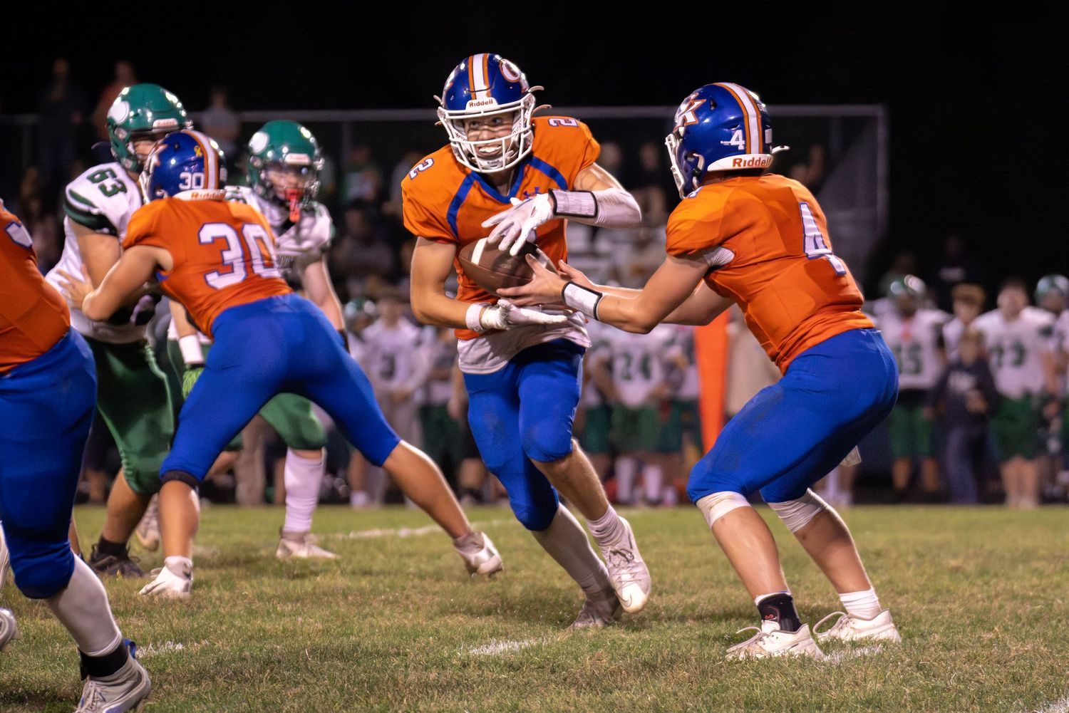 Football player in orange jersey receives handoff during nighttime high school football game.