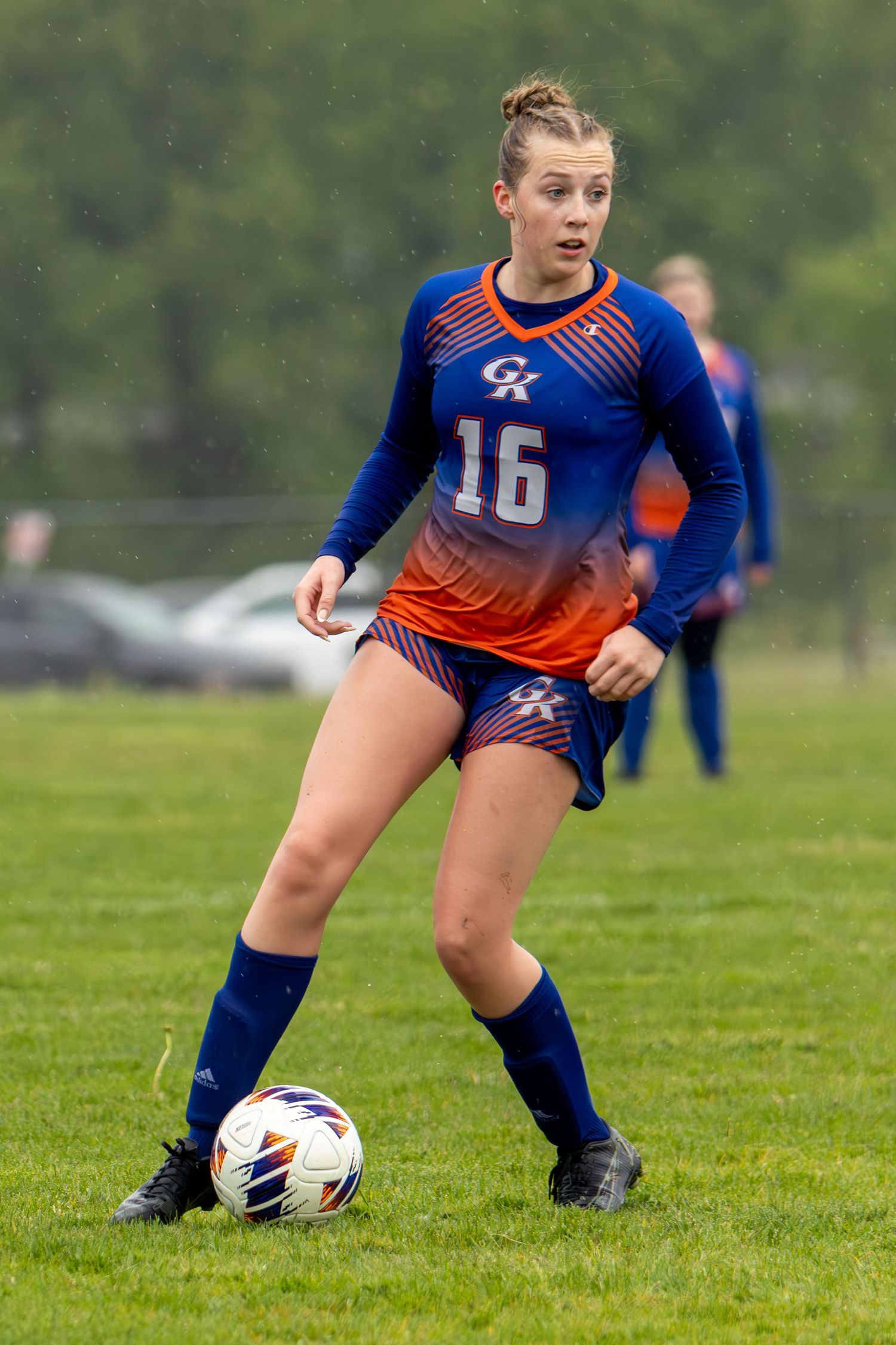 Female soccer player #16 in blue and orange uniform dribbling a ball on a green grass field during a game.