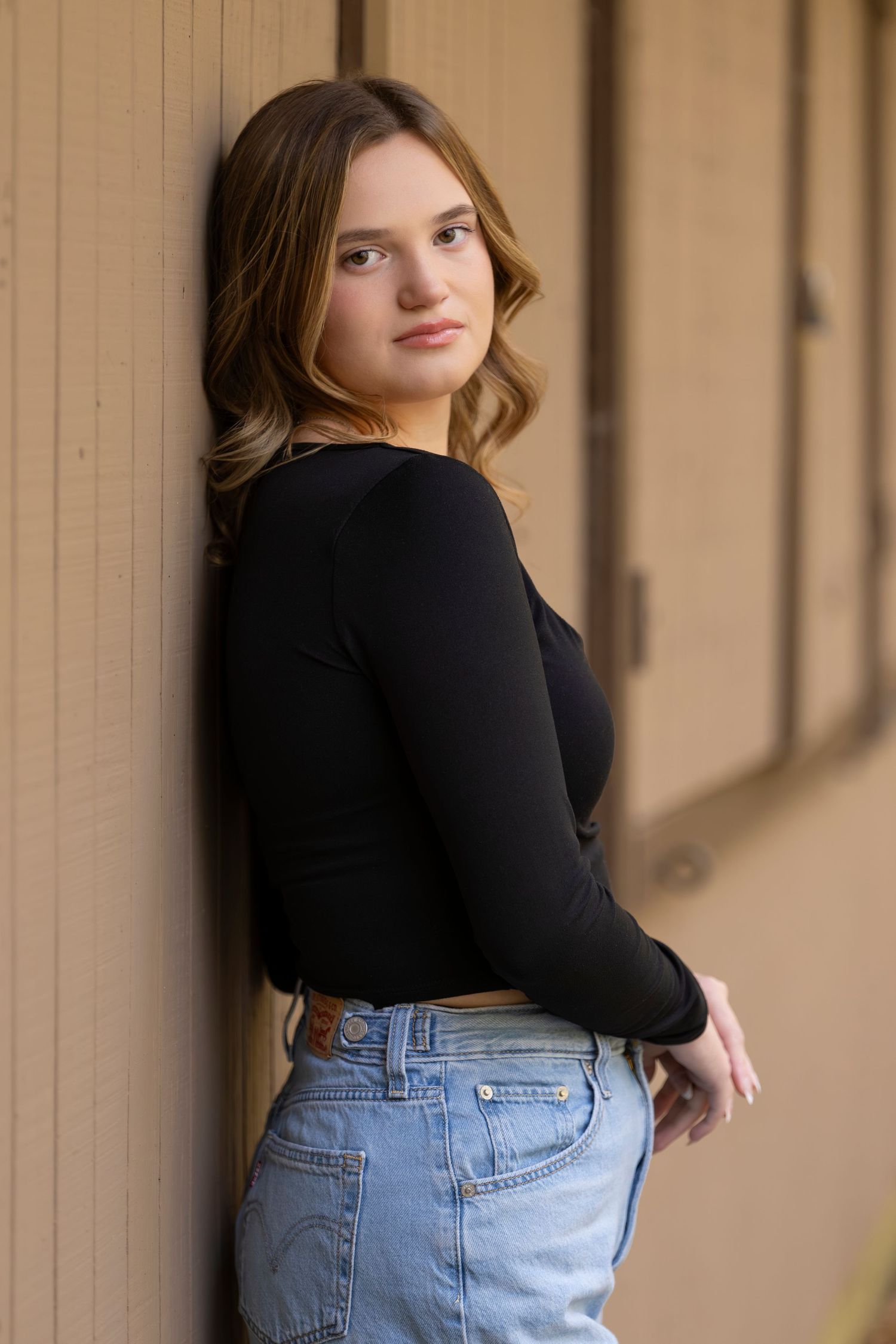 Young woman in black long-sleeve top and light blue jeans leaning against a wooden wall, posing sideways.