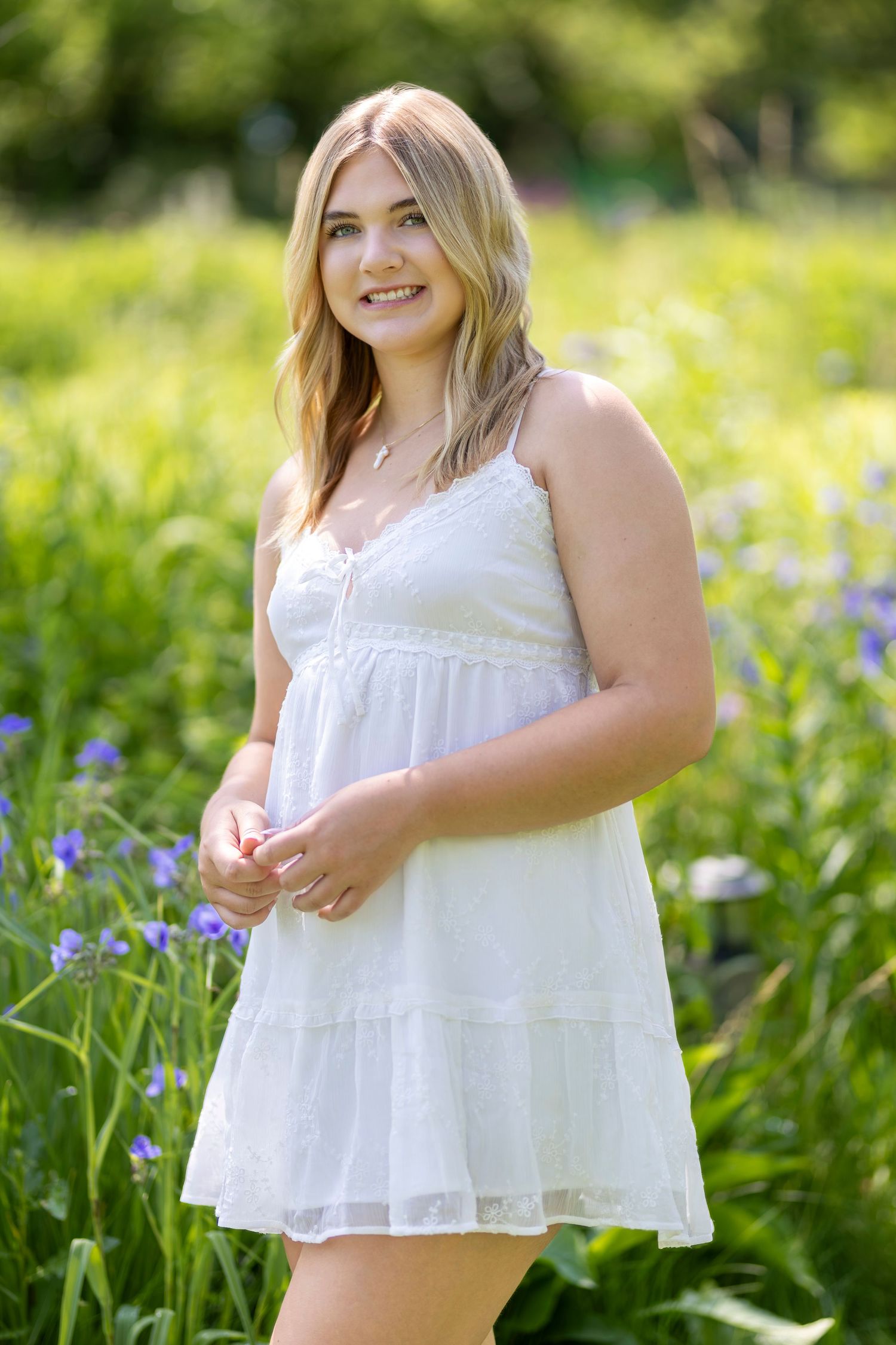 Young blonde woman in white sundress smiling outdoors among green grass and purple wildflowers on sunny day.