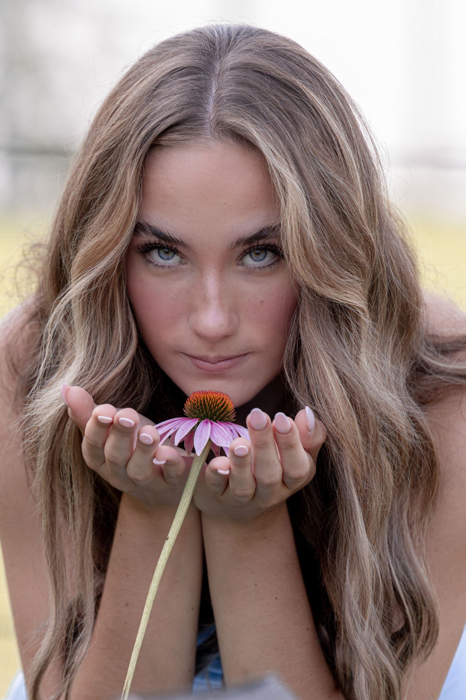 Young woman with long wavy hair holds a pink coneflower in cupped hands, gazing at camera outdoors.