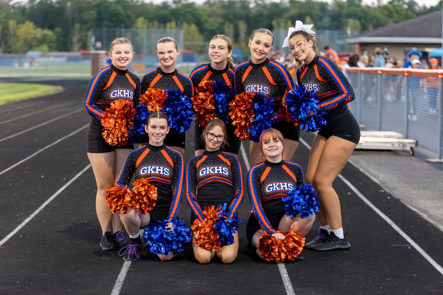 GKHS cheerleaders in blue and orange uniforms pose together on a track holding colorful pom-poms at a school event.