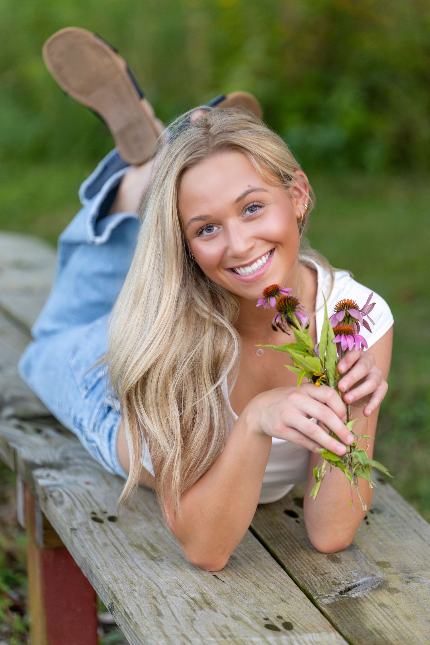 Smiling blonde woman lying on wooden bench outdoors, holding pink wildflowers, wearing jeans and white top.