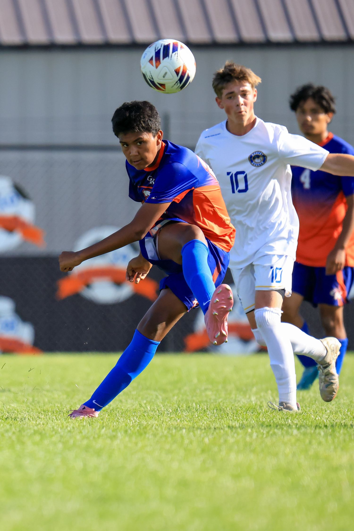 Youth soccer player in orange and blue uniform kicks ball during competitive match as opponent in white number 10 jersey defends.