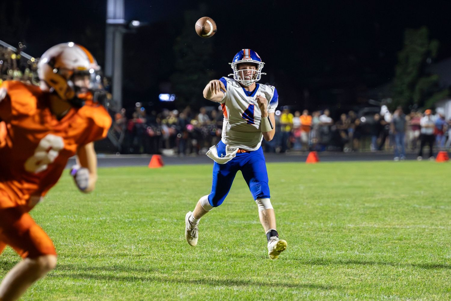 High school football player in blue uniform throwing a pass during a night game against orange-uniformed opponents.