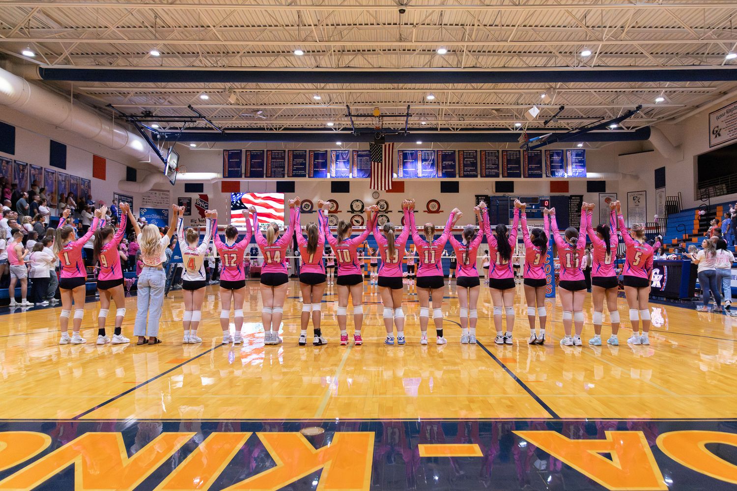 Volleyball team in pink jerseys lined up on court holding an American flag during a pre-game ceremony.