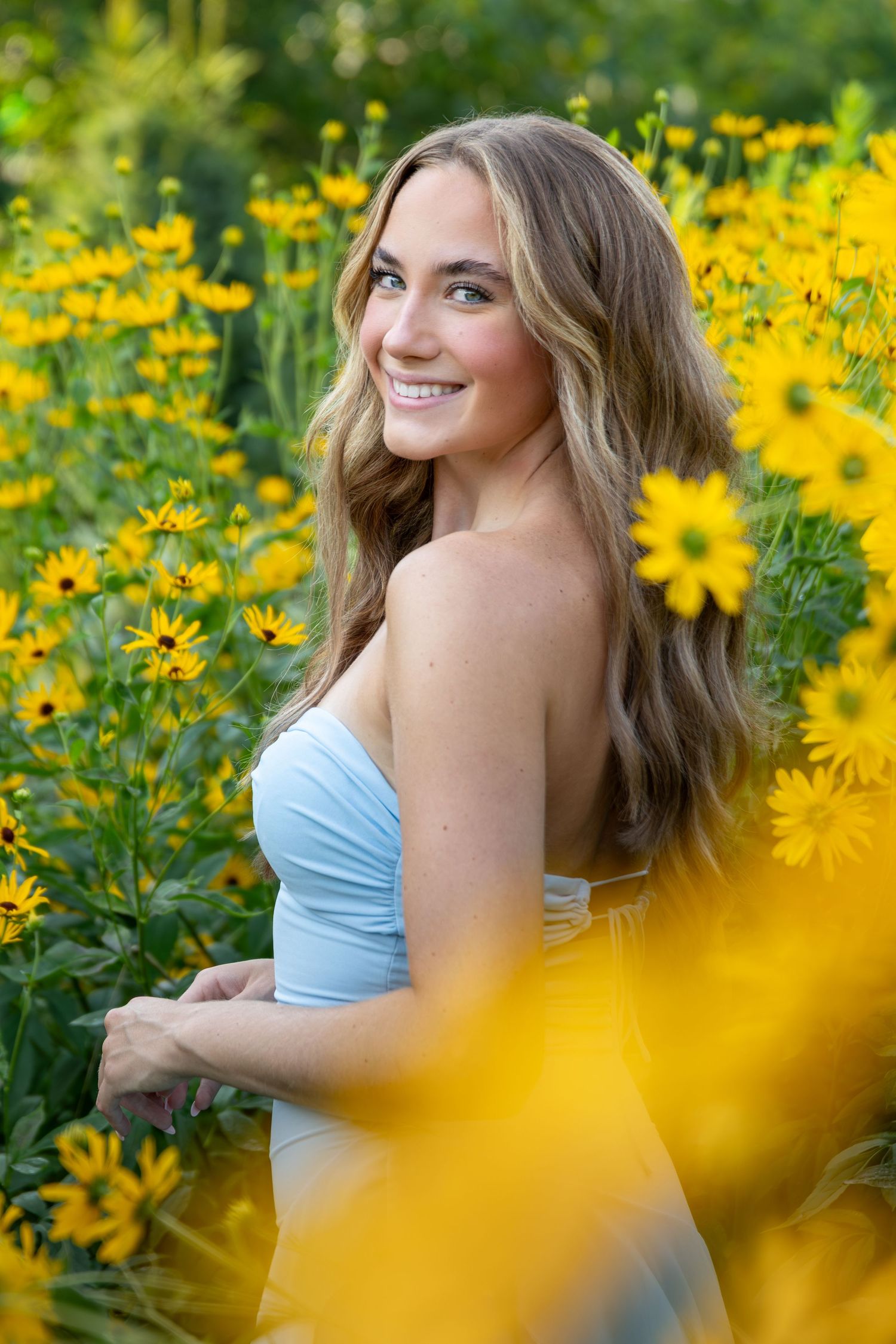 Young woman in white dress smiling among vibrant yellow wildflowers in a lush green garden setting.