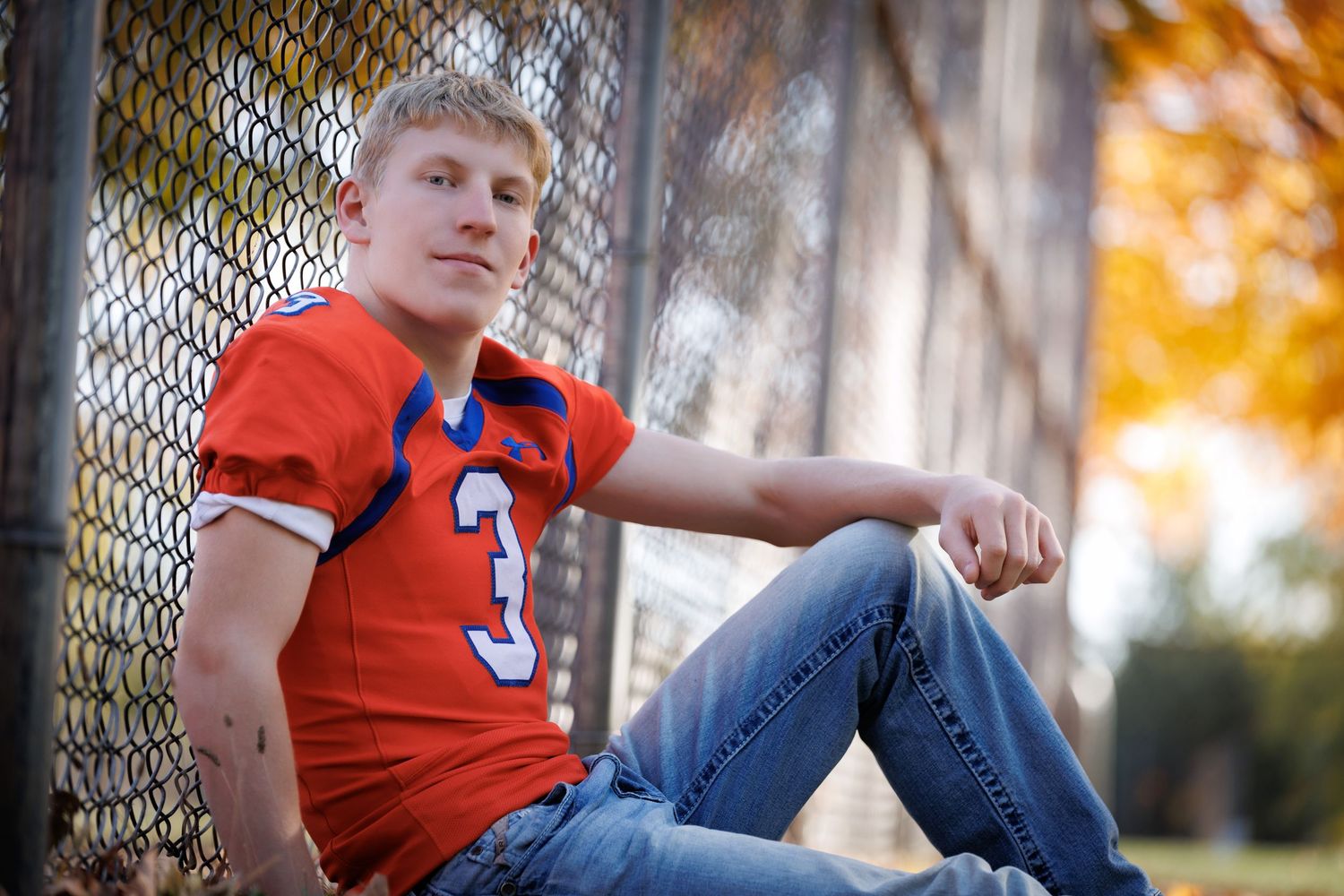 Young male athlete in orange football jersey number 3 sitting against chain-link fence outdoors.